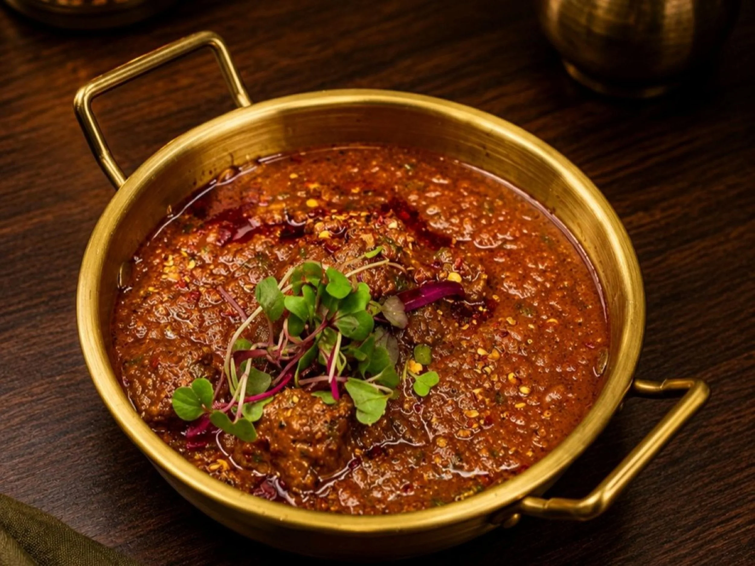 A round brass pot with a handle filled with spicy chili sauce, garnished with microgreens, on a dark wooden surface.