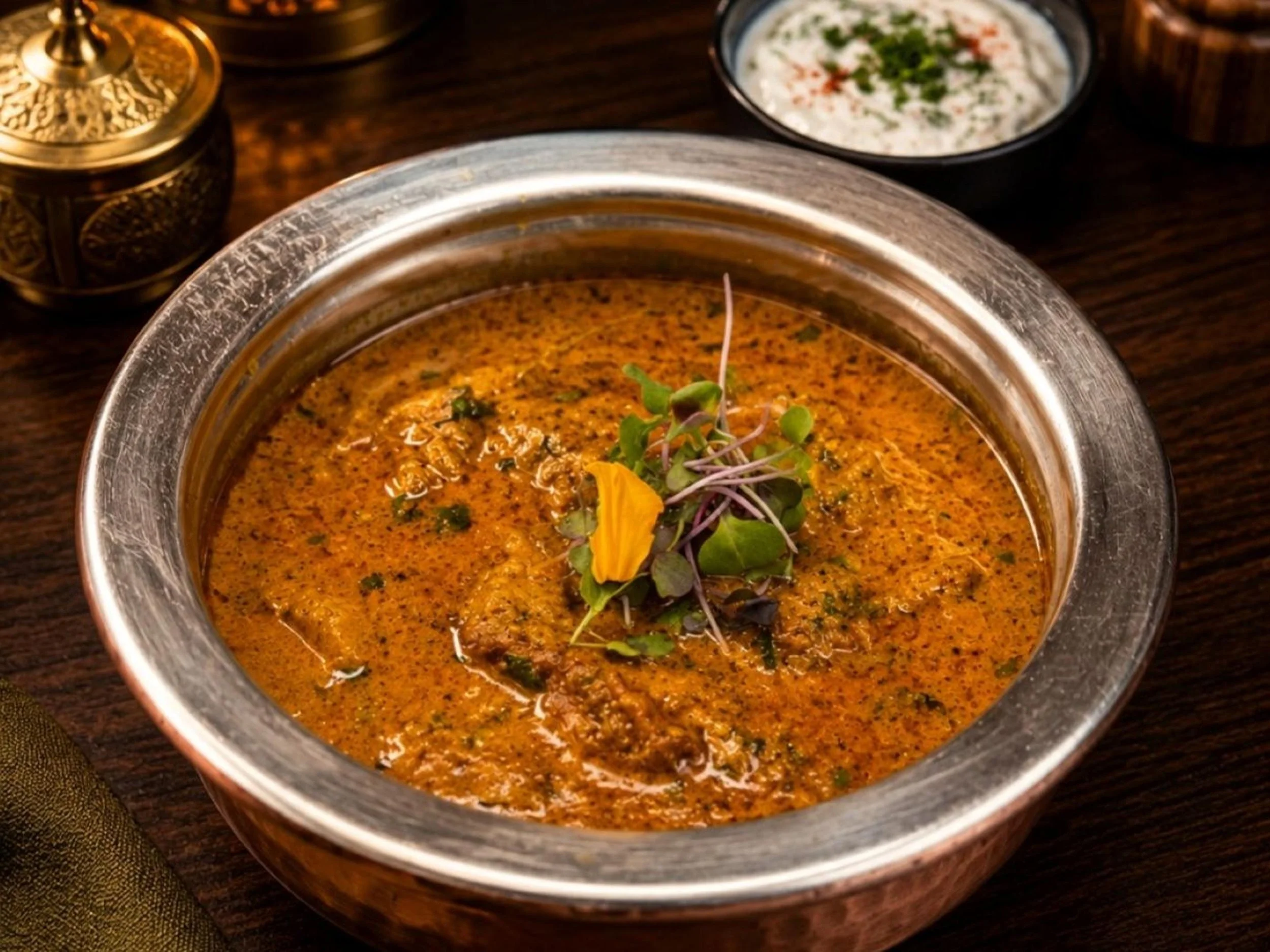 Close-up of a bowl of Indian curry garnished with microgreens and edible flower, on a wooden table with small bowls of yogurt-based raita and other condiments in the background.
