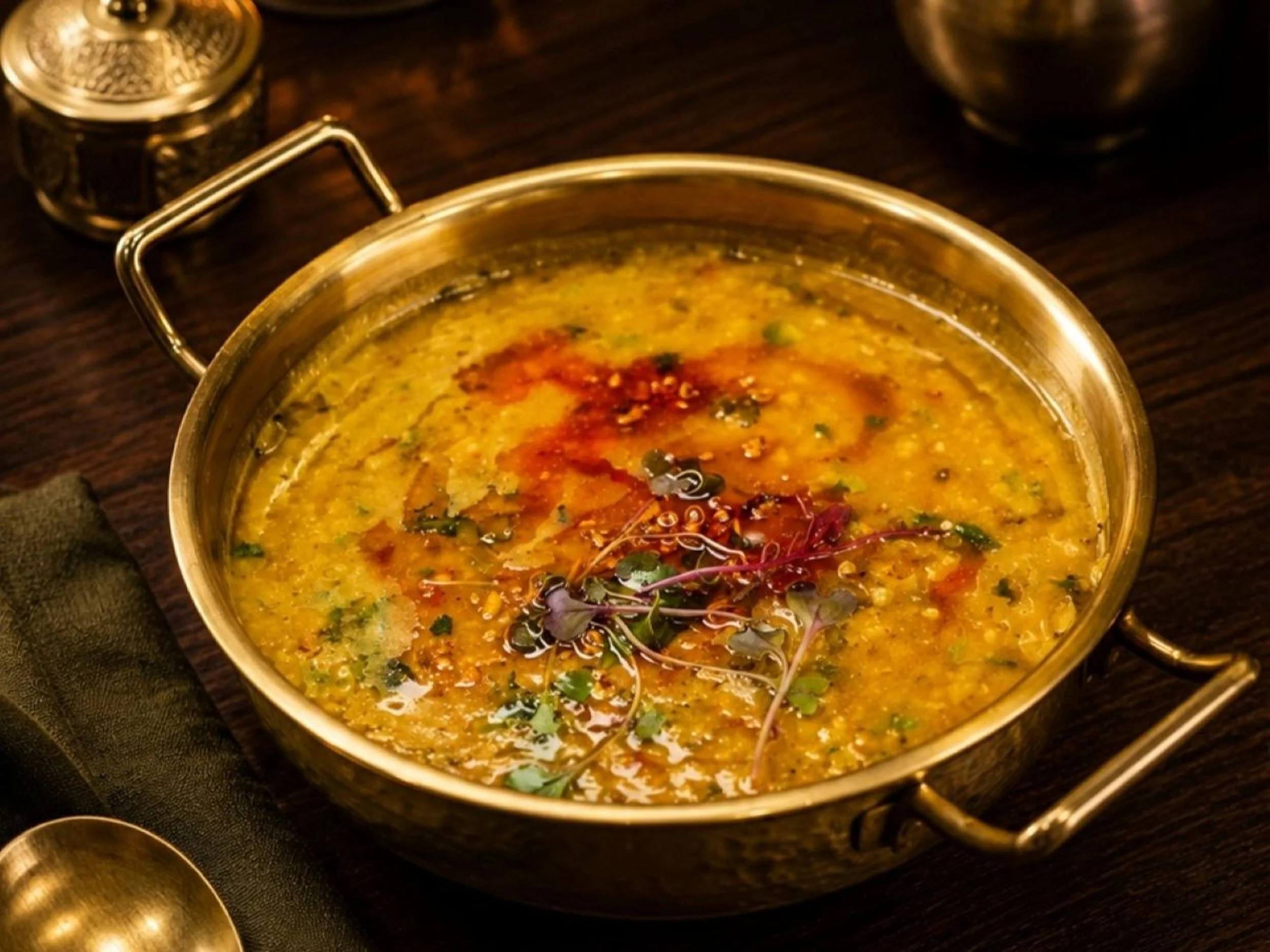 A metal pot with a yellowish-green lentil soup garnished with herbs, chili oil, and microgreens, on a dark wooden surface.