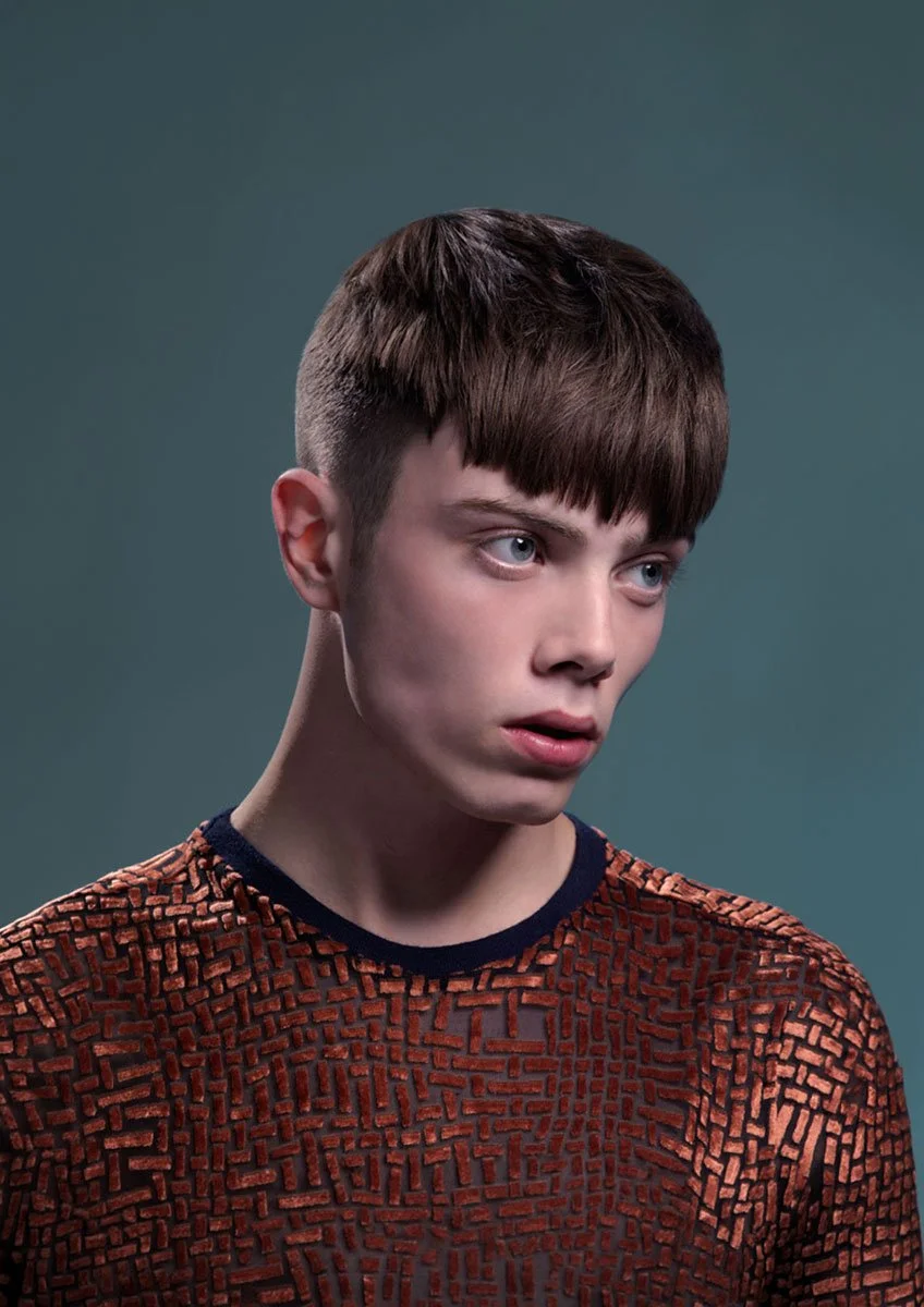 A young person with short brown hair, blue eyes, and fair skin, wearing a textured red and black patterned shirt, looking slightly to the side against a plain dark teal background.