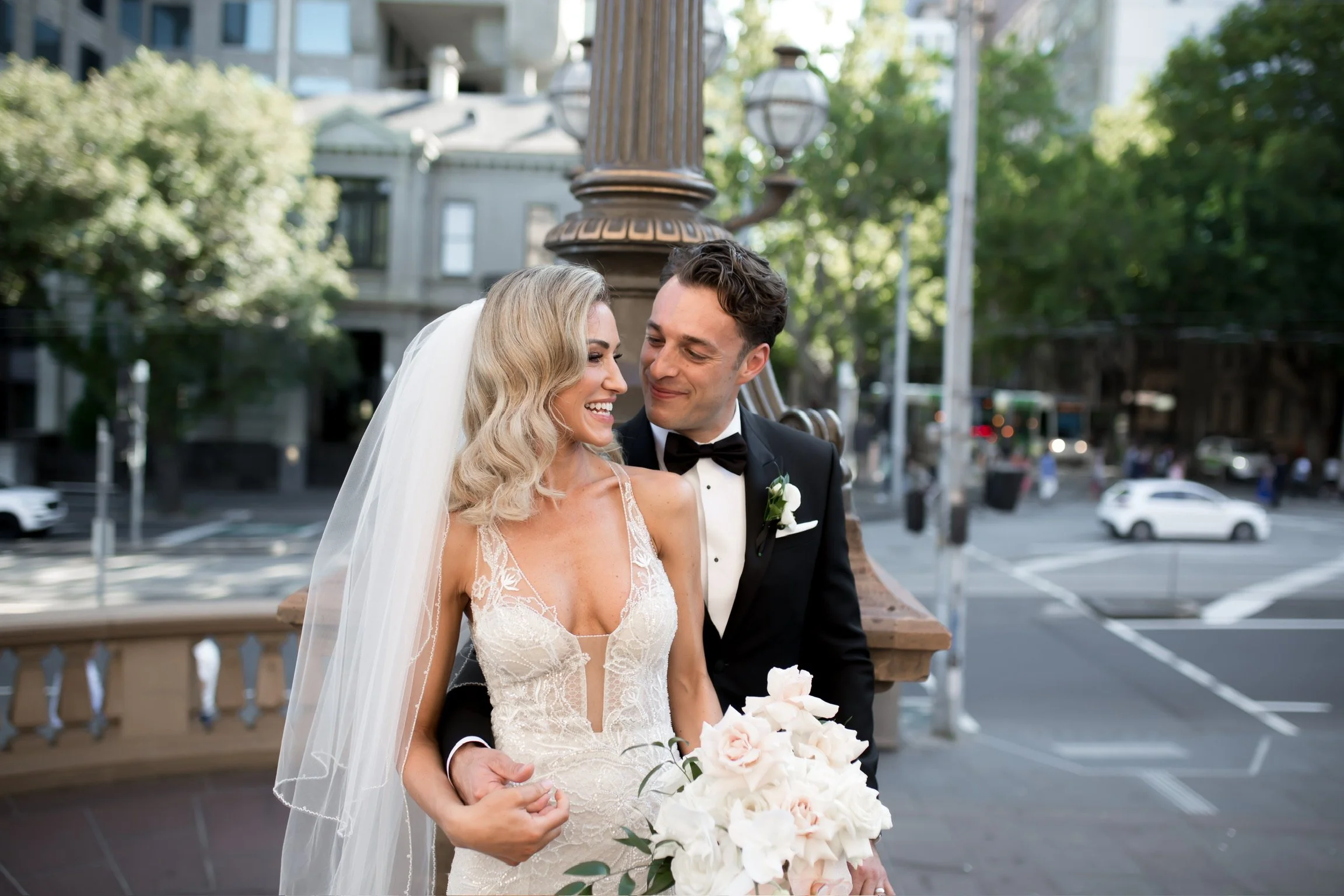 A bride and groom smiling together outdoors in a city setting. The bride is wearing an elegant lace wedding dress and a veil, holding a bouquet of white and pale pink roses. The groom is dressed in a black tuxedo with a bow tie and boutonniere. They are standing close to each other near a decorative lamppost with green trees and buildings in the background.