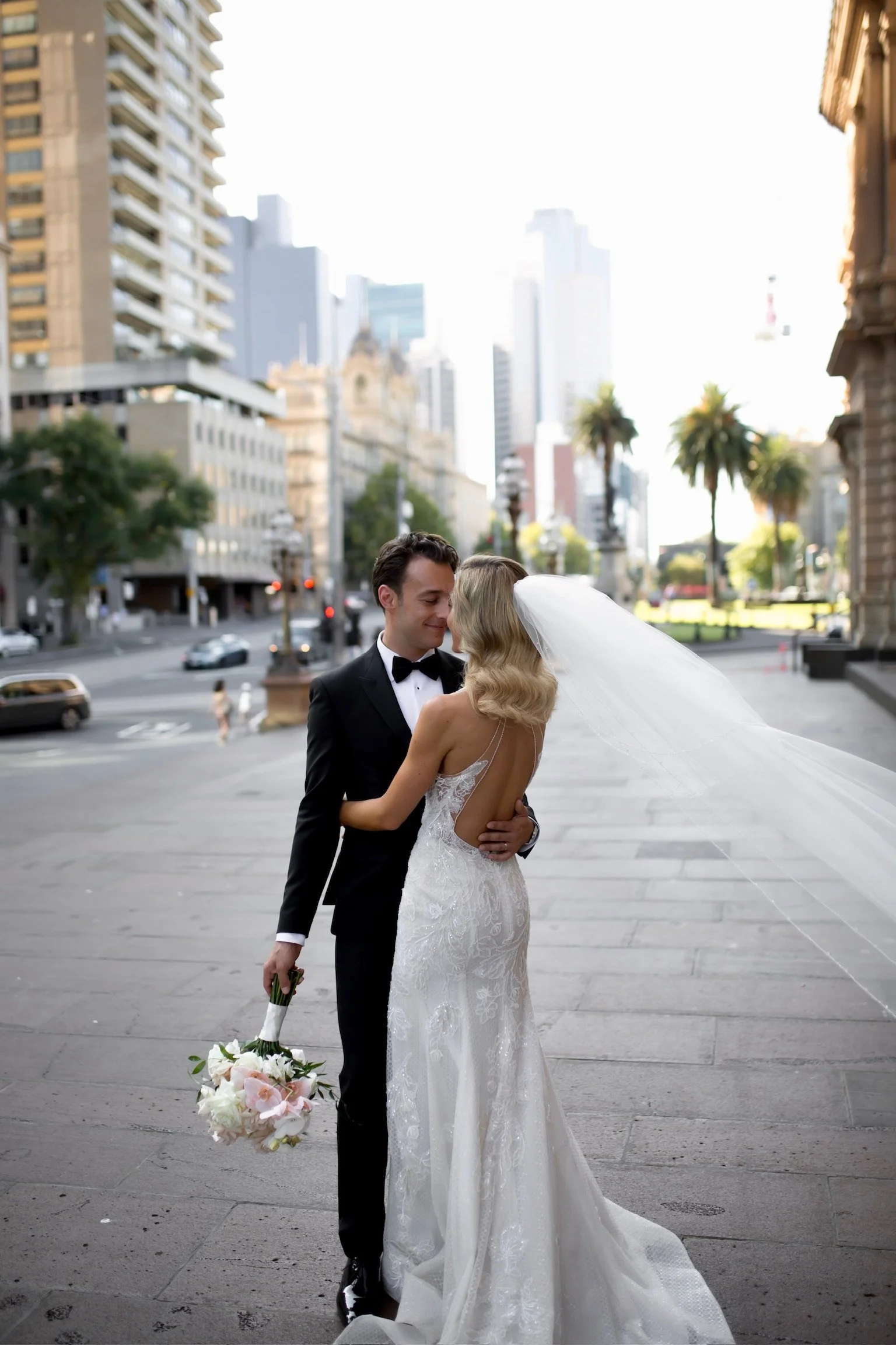 A newlywed couple sharing a close moment on a city street in the late afternoon or early evening. The groom is dressed in a tuxedo and holding a bouquet of flowers, while the bride wears a white wedding gown with a long veil. Tall buildings and palm trees are visible in the background.