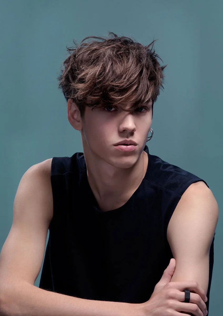 A young man with tousled brown hair, wearing a sleeveless black shirt, looking serious against a muted green background.