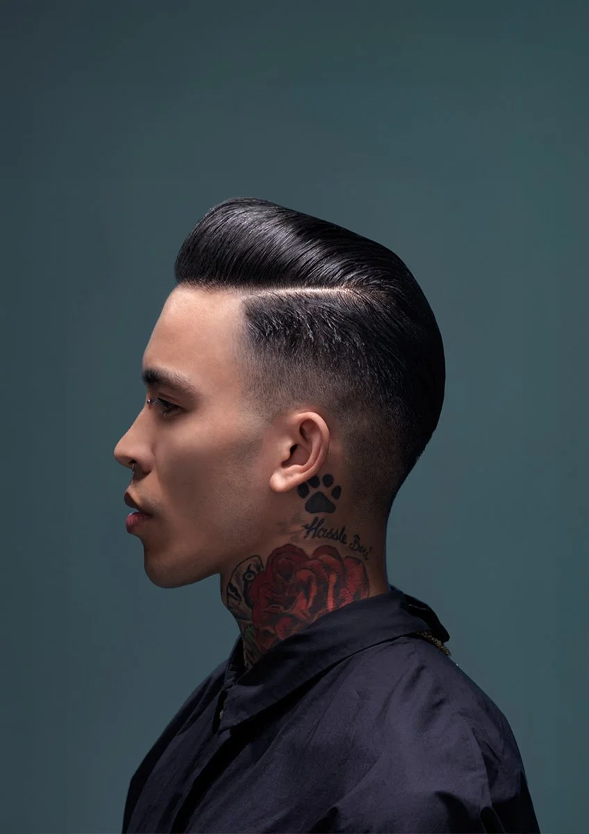 Side profile of a young man with slicked-back black hair, tattoos on his neck including a red rose and paw print, wearing a dark shirt against a plain background.