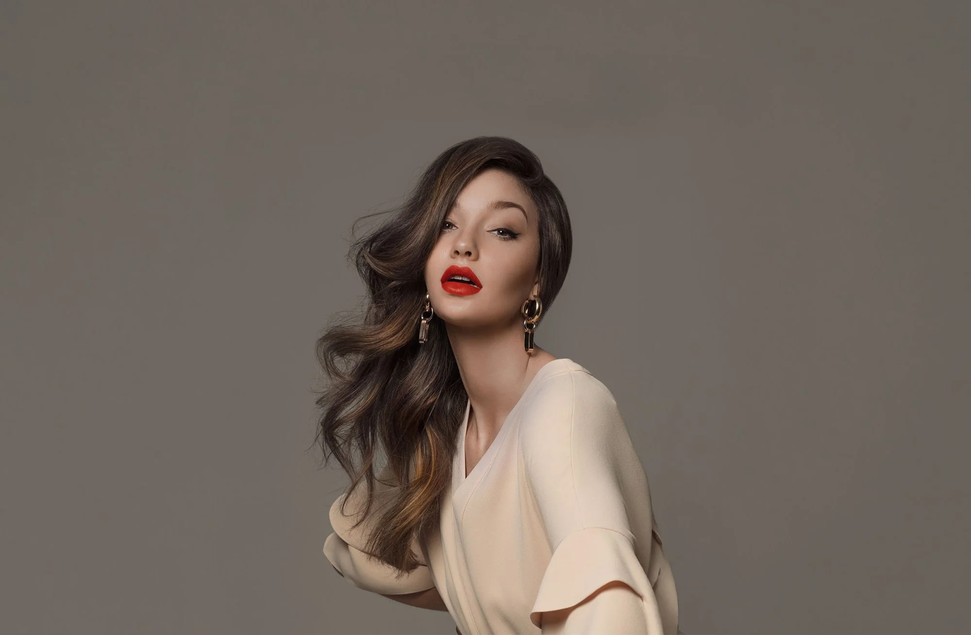 A woman with long, wavy brown hair and red lipstick poses against a gray background. She is wearing earrings and a cream-colored blouse with a slight V-neck.