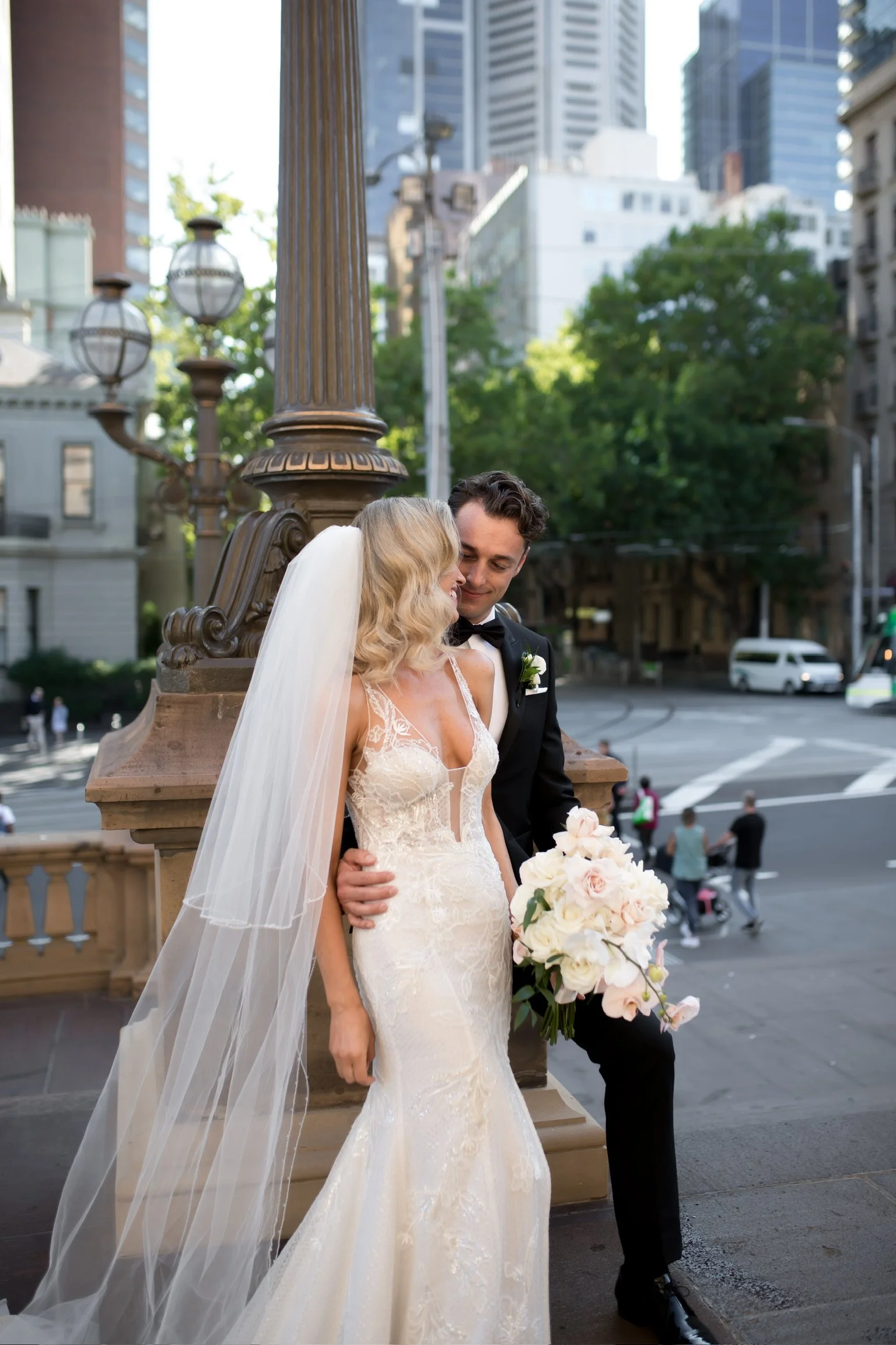 A bride and groom sharing a kiss on a city street. The bride is wearing a lace wedding gown with a veil, holding a bouquet of pink and white roses. The groom is in a black tuxedo with a bow tie. There are tall buildings, trees, and streetlights in the background, along with some pedestrians and a vehicle passing by.
