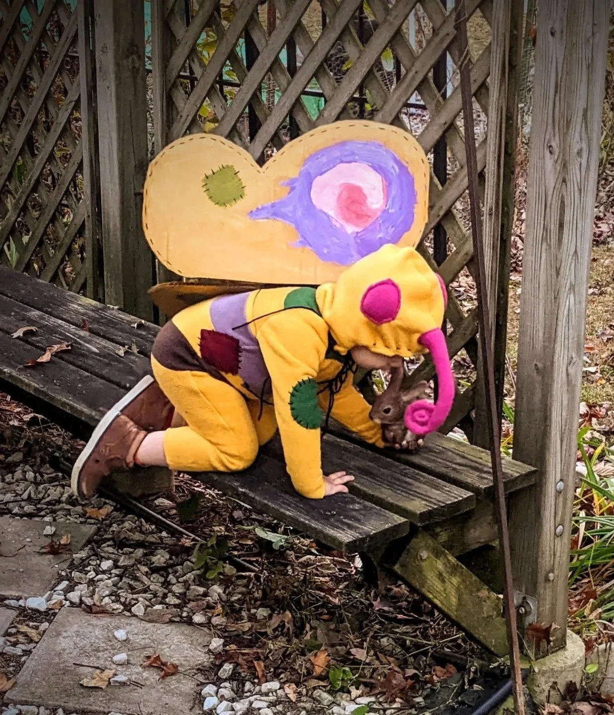 Child dressed in a yellow butterfly costume with pink and green spots interacting with a small brown rabbit on a wooden bench outdoors, with a painted backdrop of a heart and a swirl design based on the artwork "Glimmer Bairn" by Nils Hamm