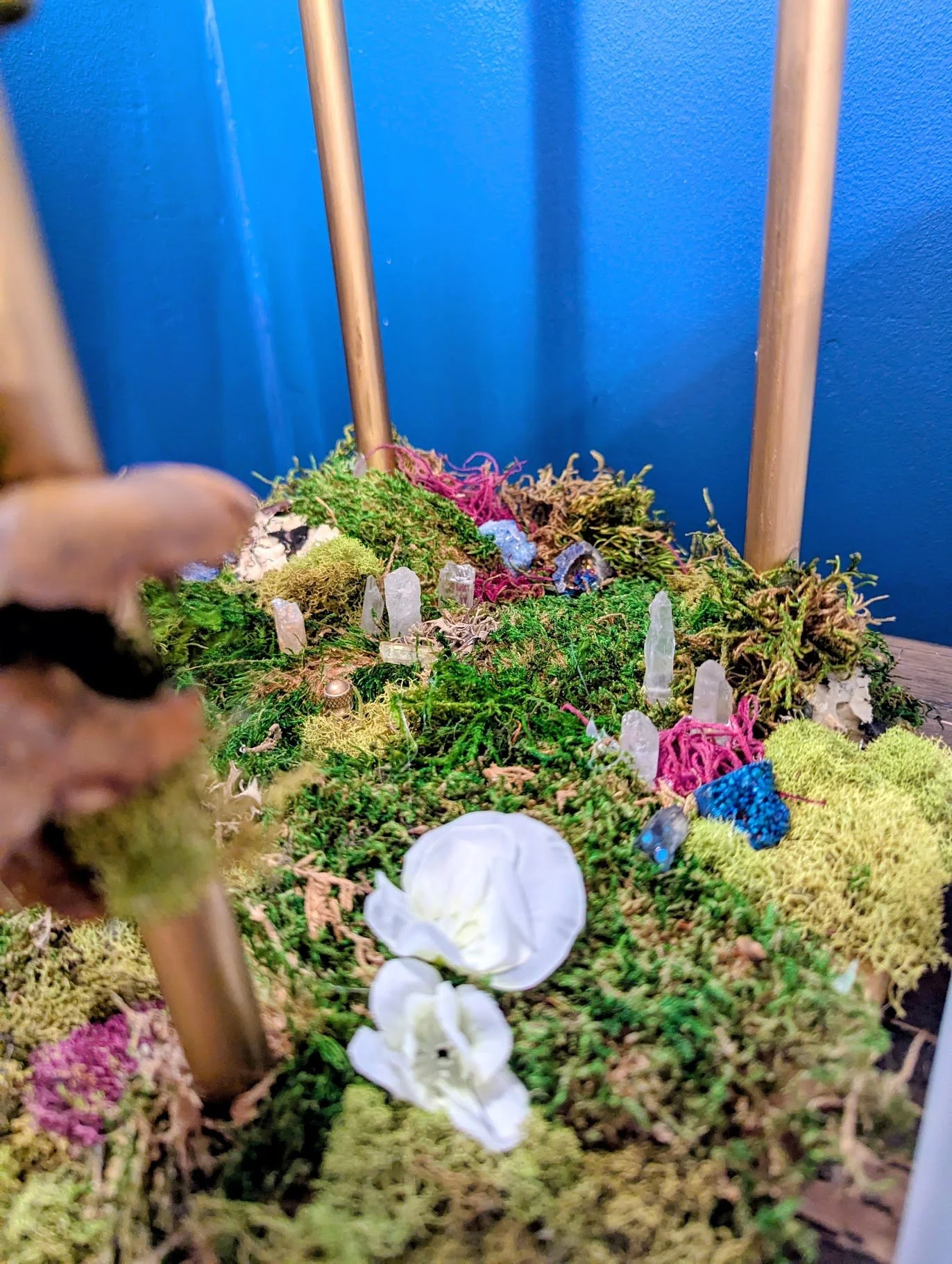 Miniature scene made on base of lamp with moss, small crystals, and white flowers on a mossy landscape with wooden sticks and a blue background.