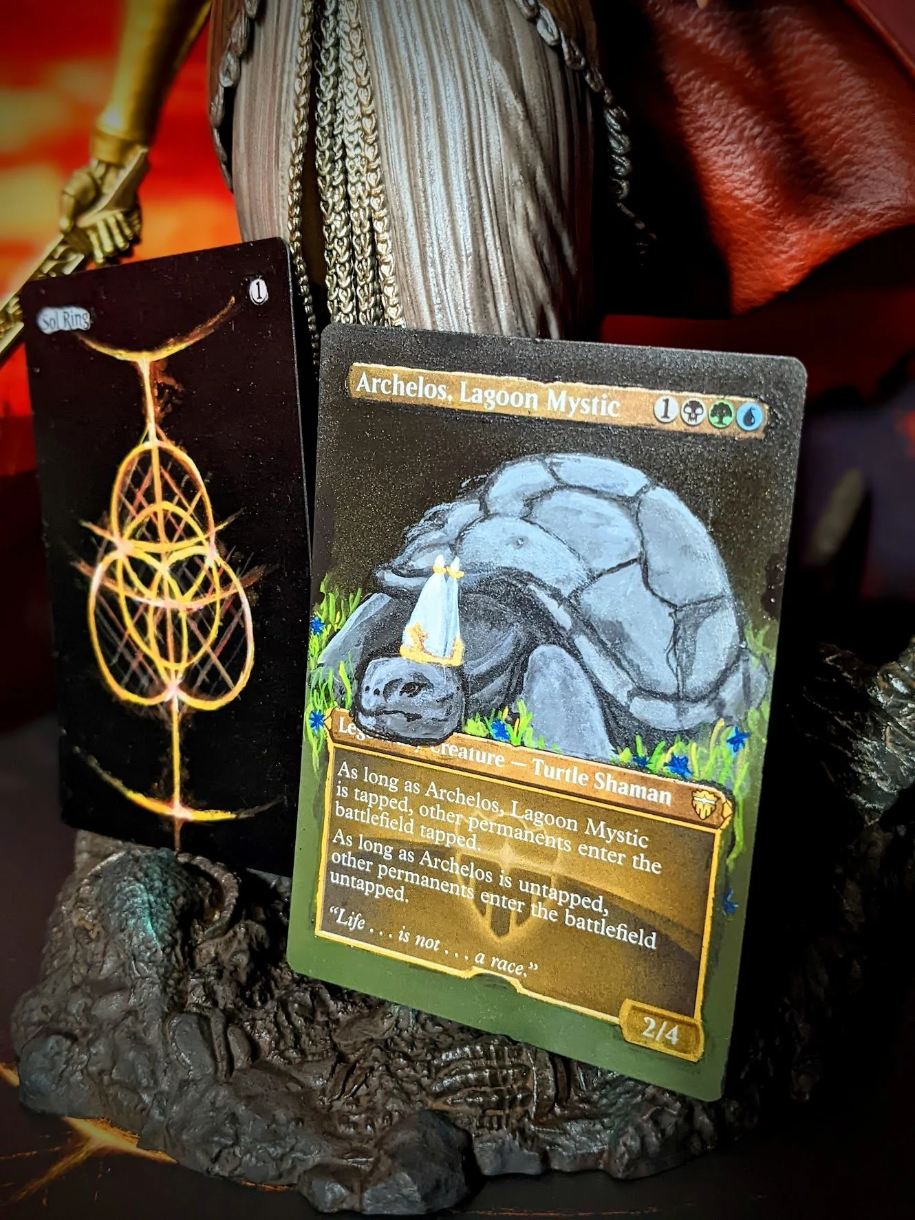 Magic:  card named 'Archelos. Lagoon Mystic' showing a turtle creature, and another card with a symbol called 'Sol Ring'. The cards are placed on a textured, possibly volcanic or rocky surface, with a background of other objects and a reddish cloth.