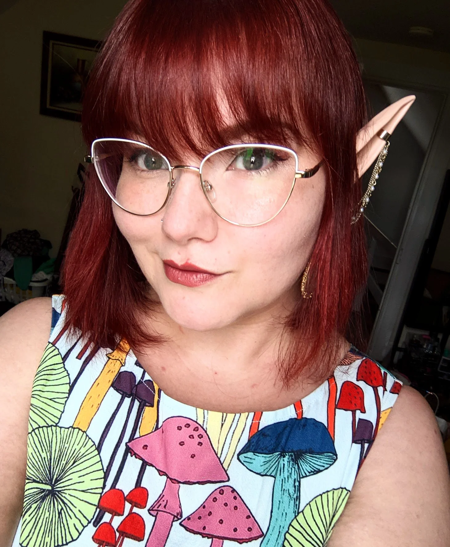 A woman with red hair, wearing glasses with a decorative chain on the temple, and elf-like pointed ear accessories. She is smiling and wearing a colorful dress with a mushroom and umbrella pattern.