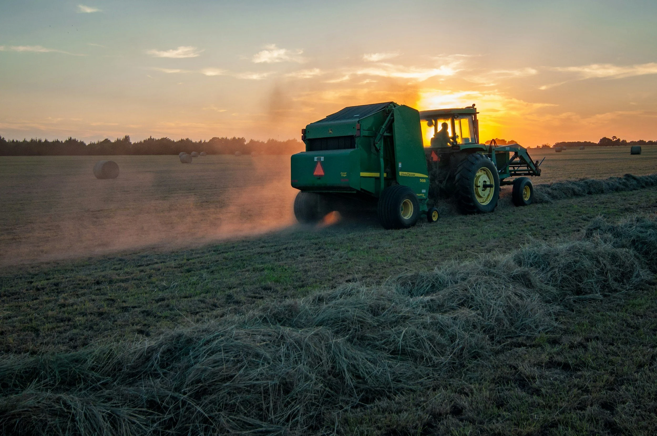 A green John Deere tractor operating in a field at sunset with hay bales scattered across the landscape.