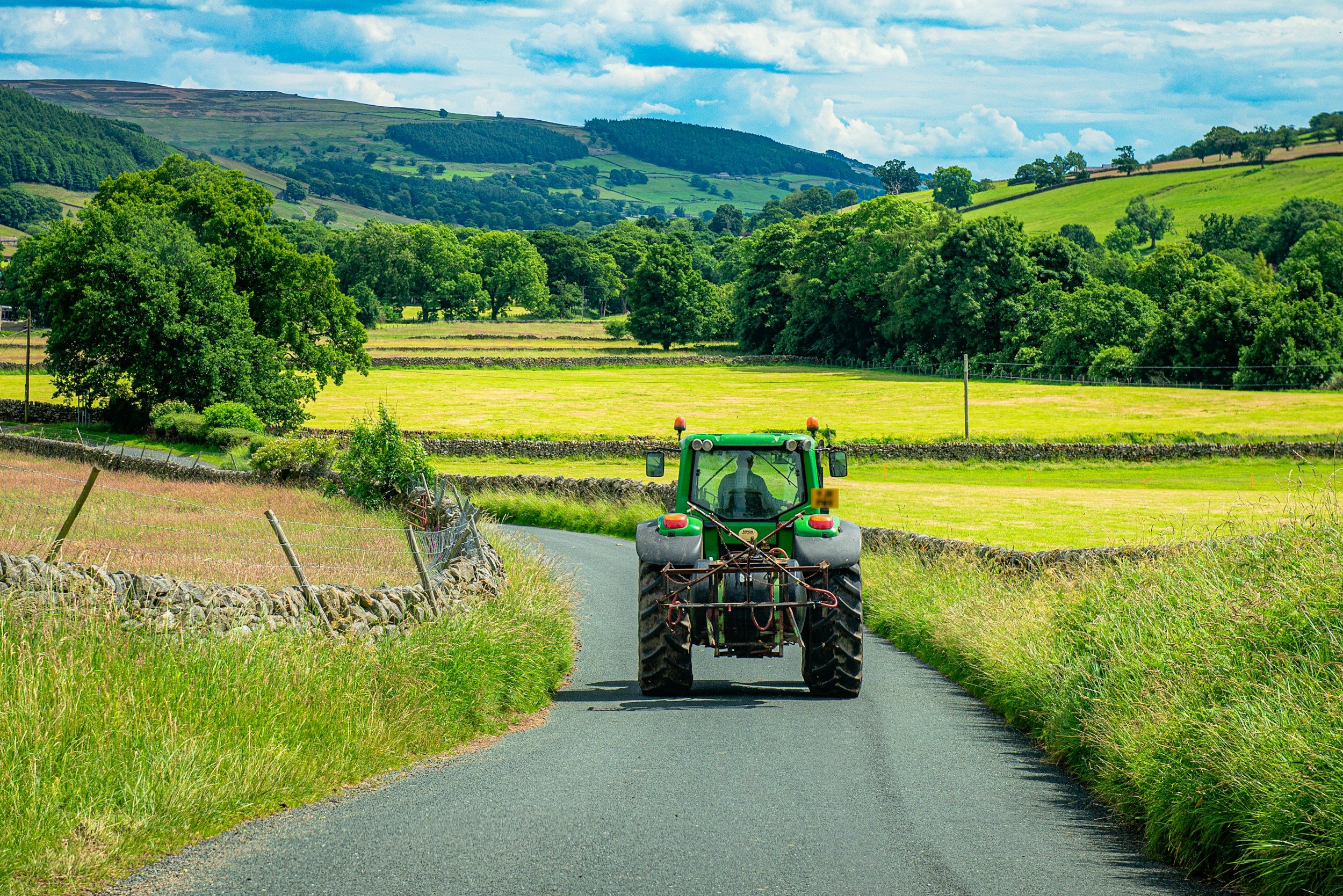 A tractor driving down a narrow rural road surrounded by green fields and trees, with rolling hills and a partly cloudy sky in the background.