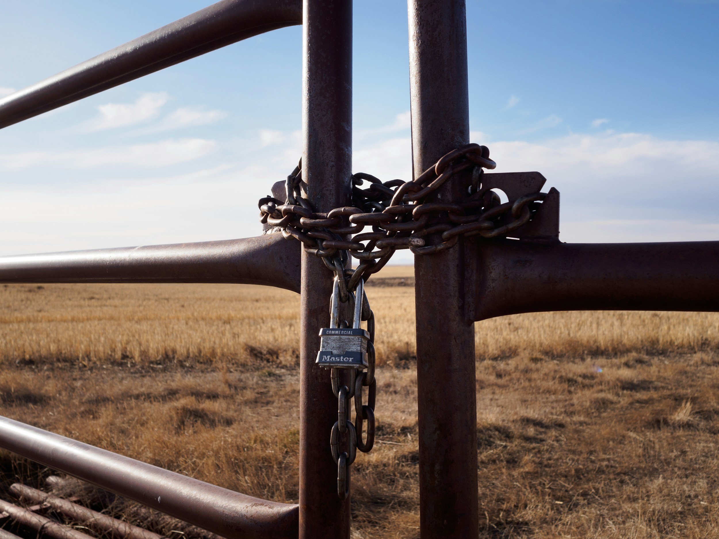 A close-up view of a metal gate with a chain and padlock securing it, set in an open field with a clear blue sky.