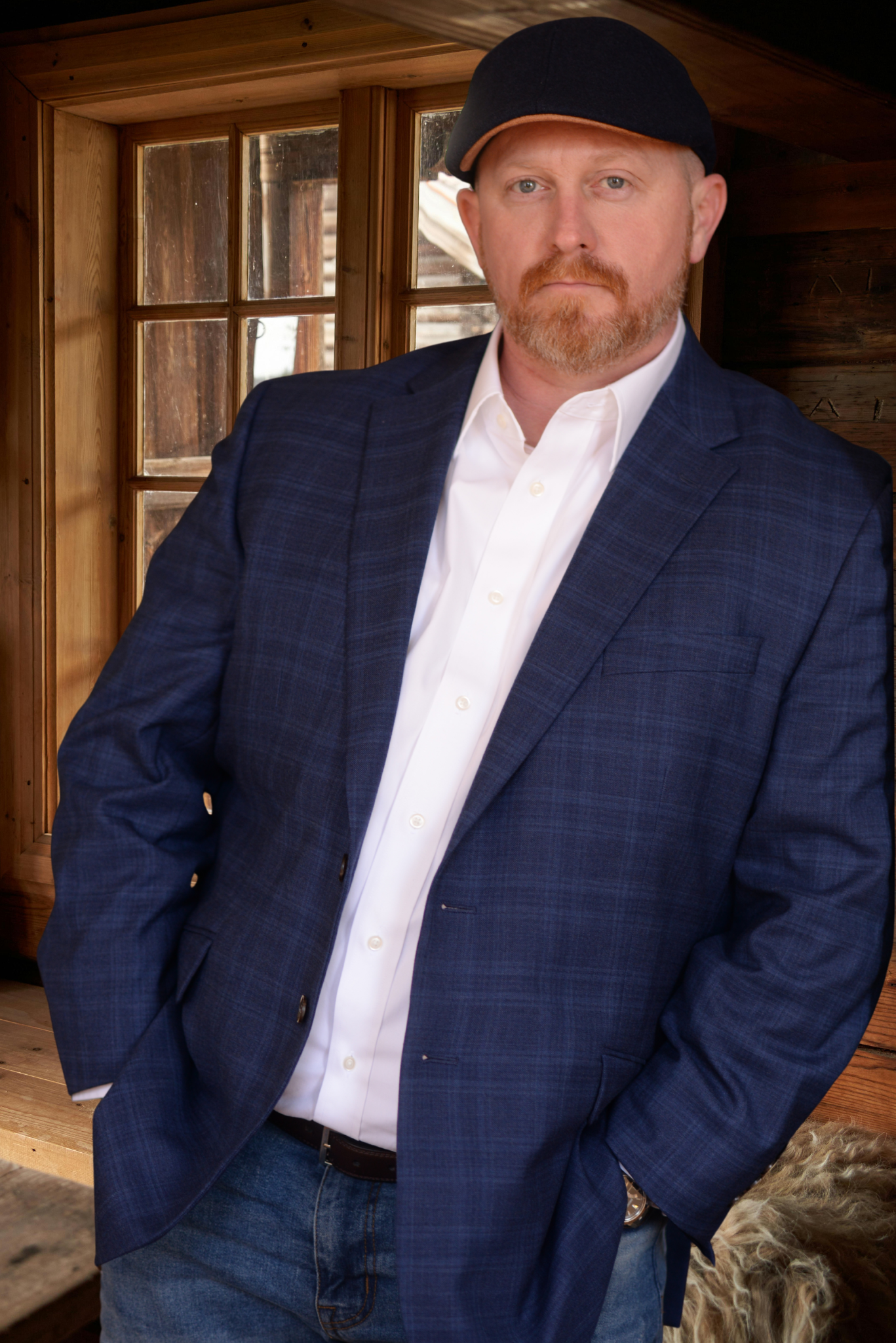 A man with light skin, a reddish beard, and blue eyes wearing a dark blue blazer, white button-up shirt, and a black cap with a tan bill standing in a wooden room with a window behind him.