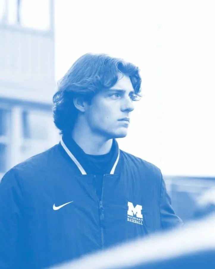 Young man with wavy hair wearing a black Nike jacket with University of Michigan baseball logo, standing indoors with natural light.