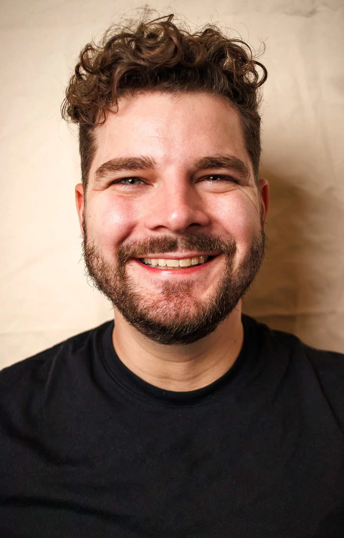 About page of Myles Travitz. A close up portrait of him with curly brown hair and beard, wearing a black shirt, against a light background. Learn more about his journey with pottery!