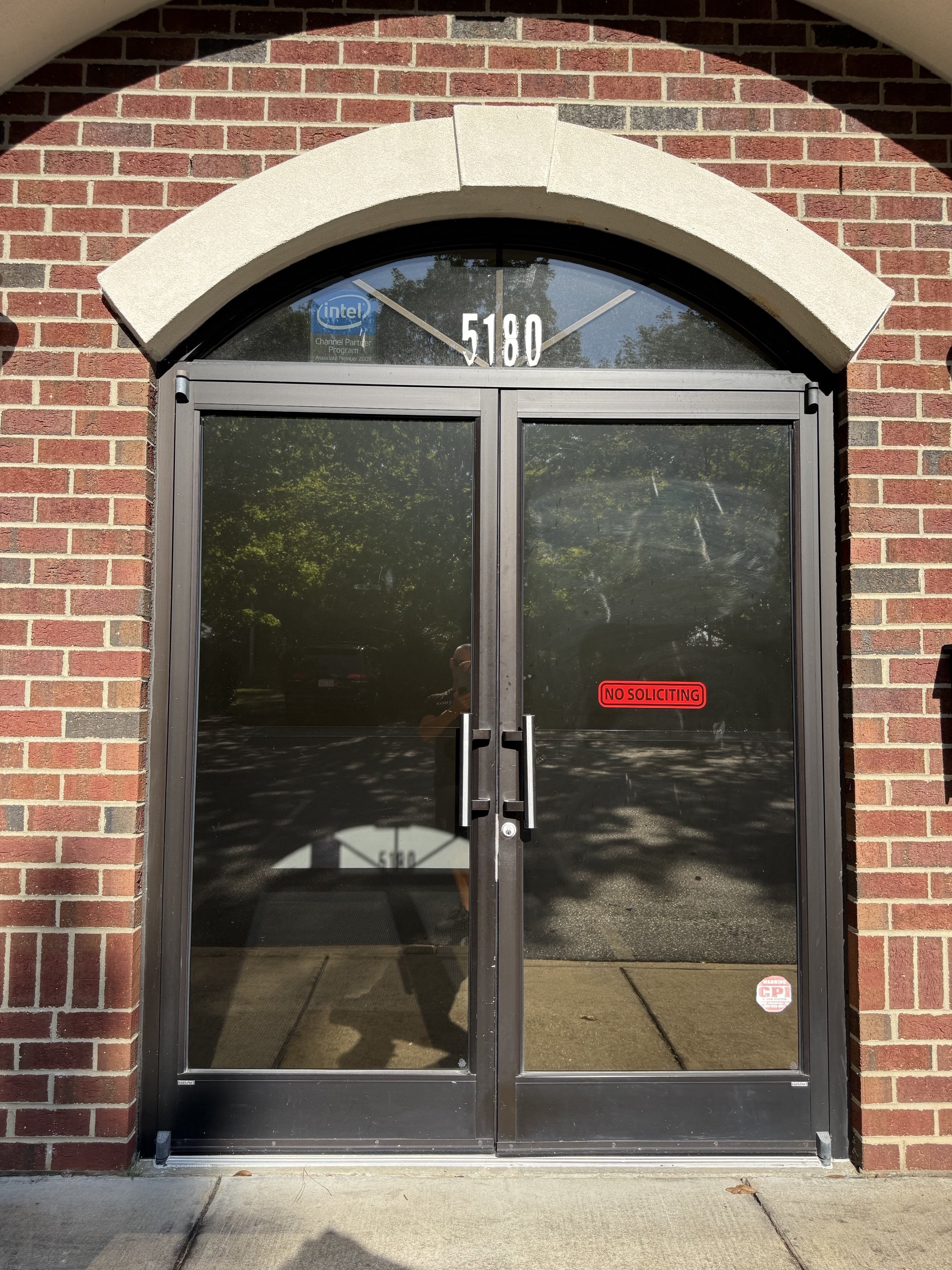 A glass door with a brick arch above it, displaying the number 5180 at the top and a red 'No Soliciting' sign on the right door panel. The reflection of trees and a person can be seen in the glass.
