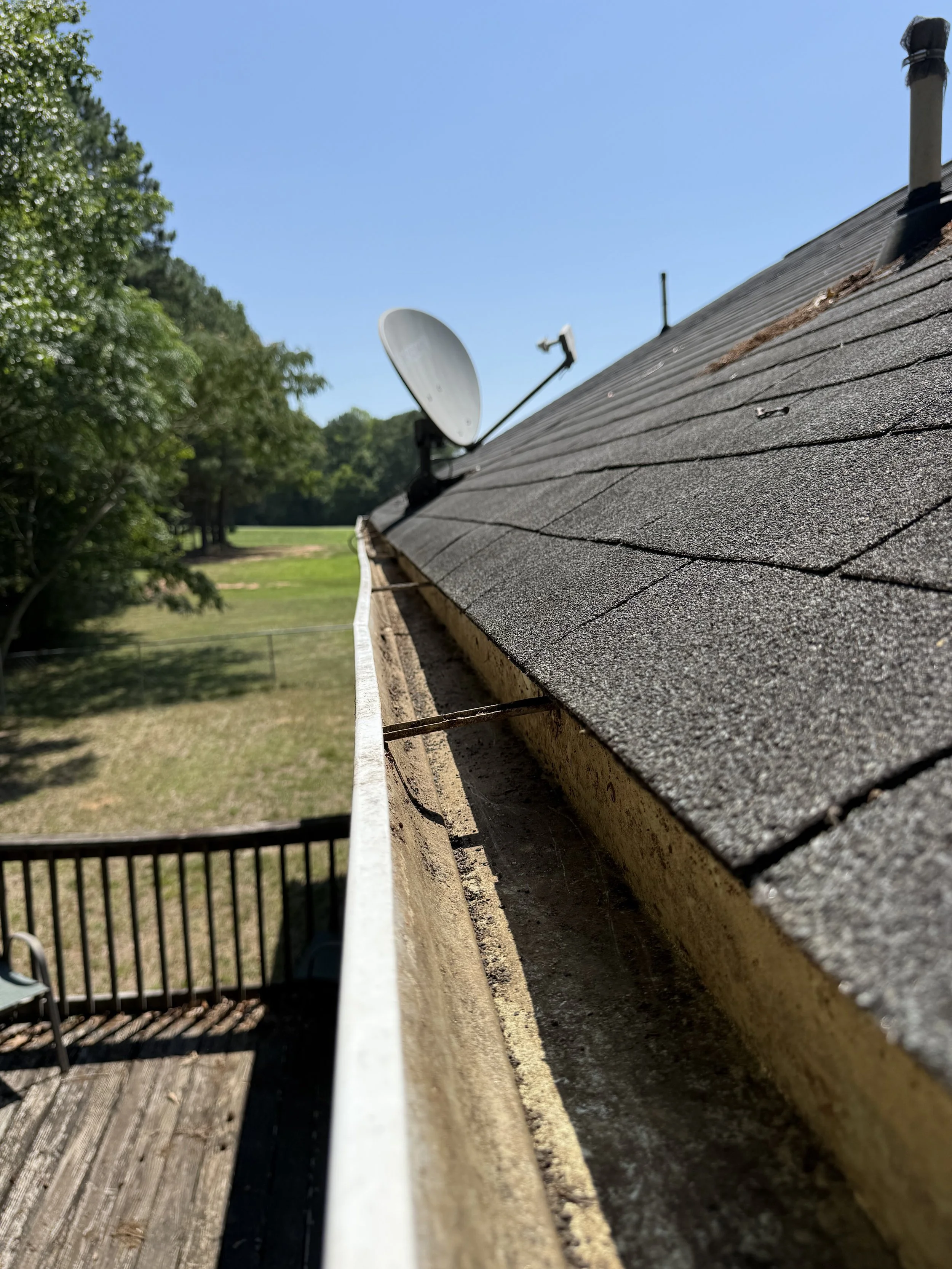 View of a roof covered with asphalt shingles, with a satellite dish and a vent pipe, against a clear blue sky and green trees in the background. Gutter cleaned of all debris.