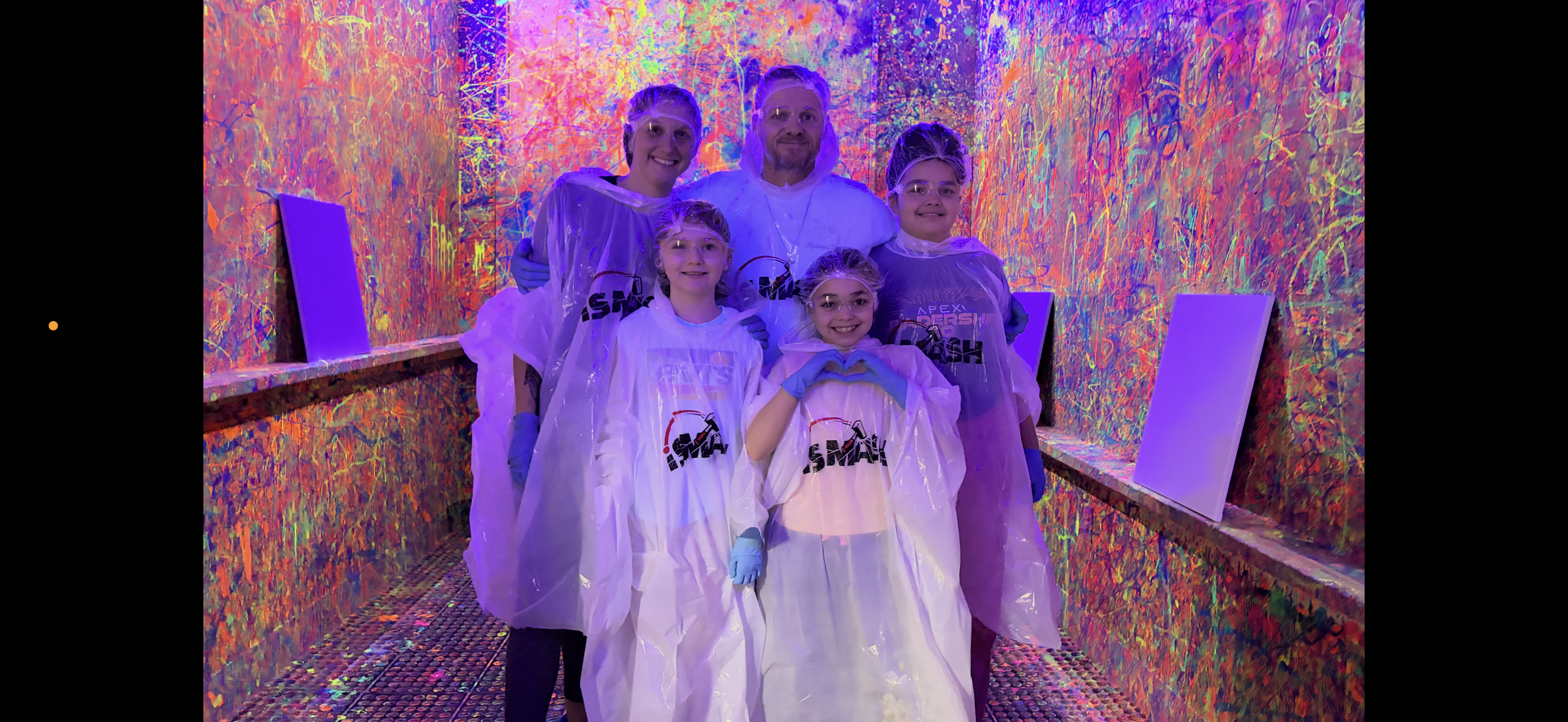 Group of five, the owner and his family in protective coveralls and glasses, standing in a colorful, illuminated art installation with neon paint splatters on the walls and floor.
