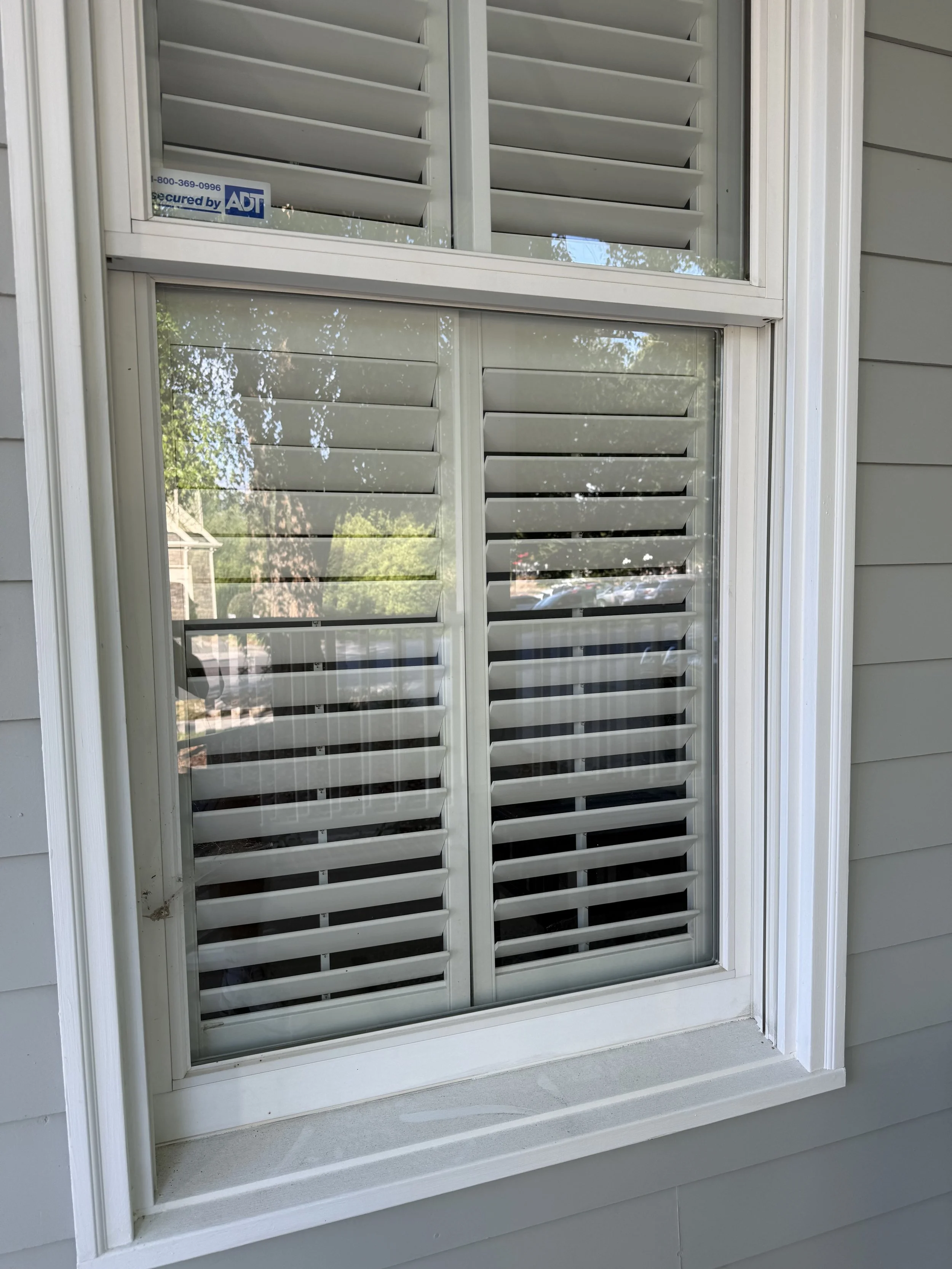 A  very dirty window with white shutters on a house with the window frame very dirty.