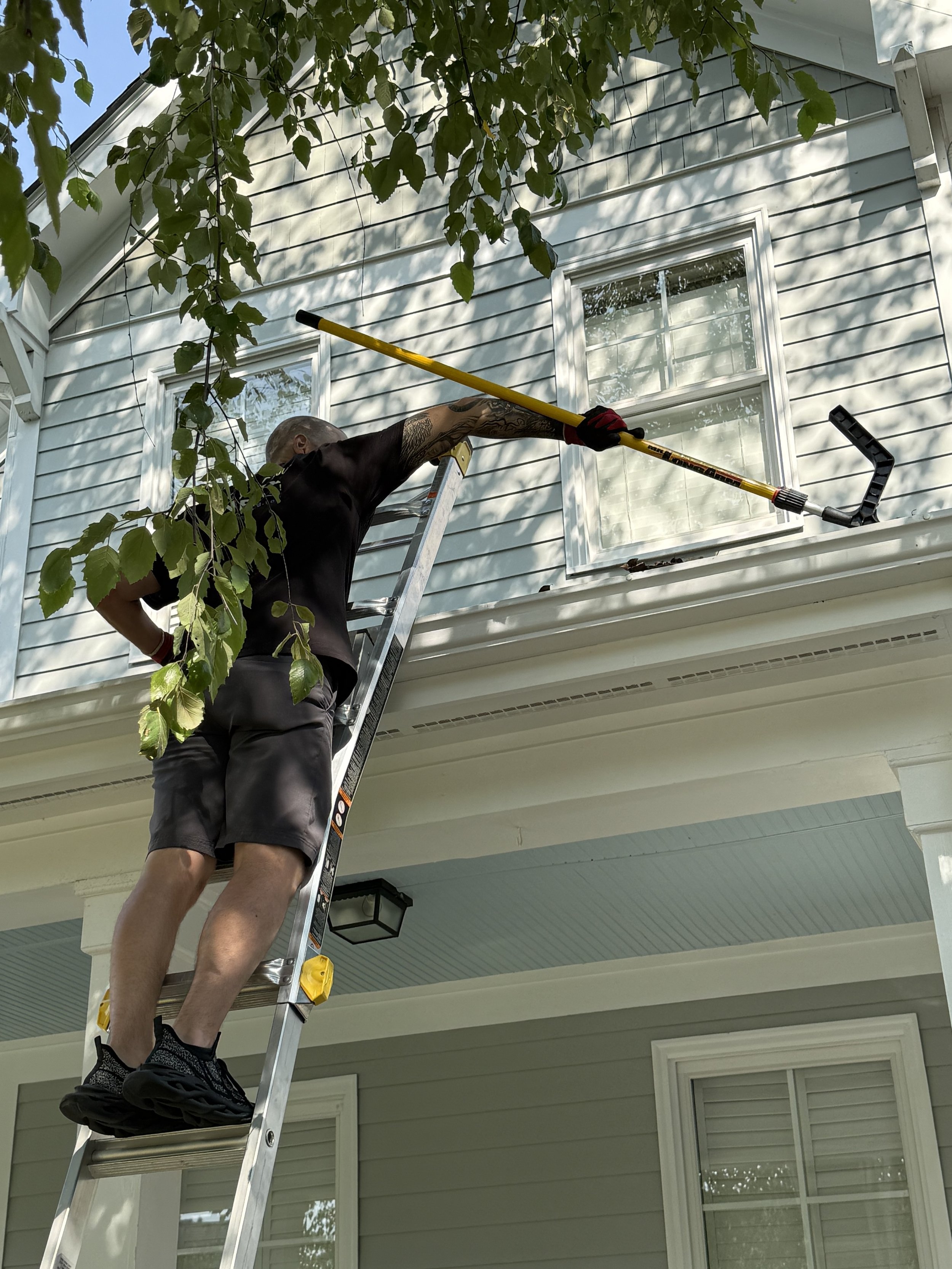 Person cleaning or washing windows on the upper floor of a light-colored house using an extension tool, standing on a ladder.