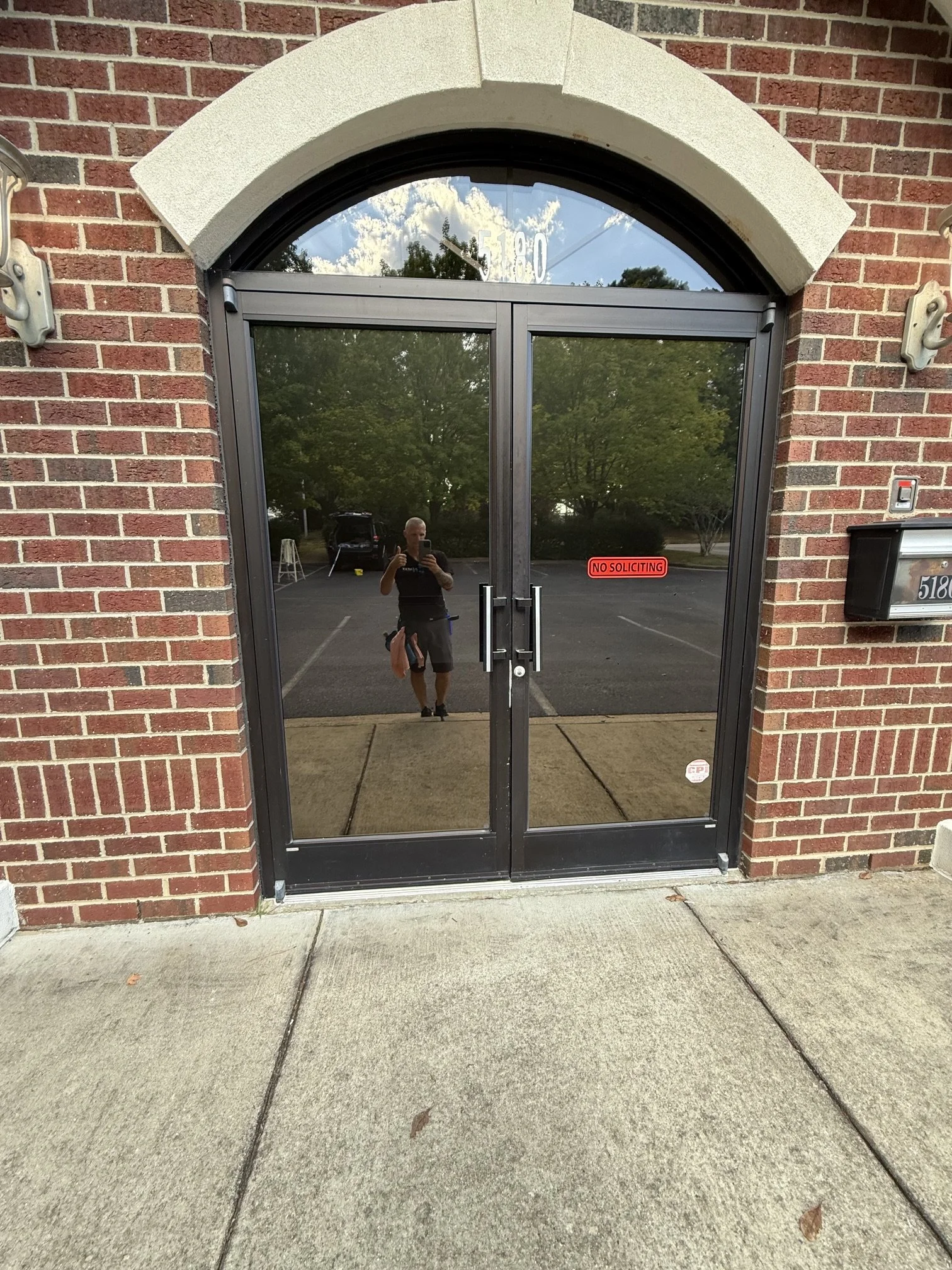 The image shows a glass door with a brick wall around it. There is a person visible in the reflection taking a photo, and they are giving a thumbs-up. The door has a sign that reads 'NO SOLICITING' and the address number '5180' is visible above the door.