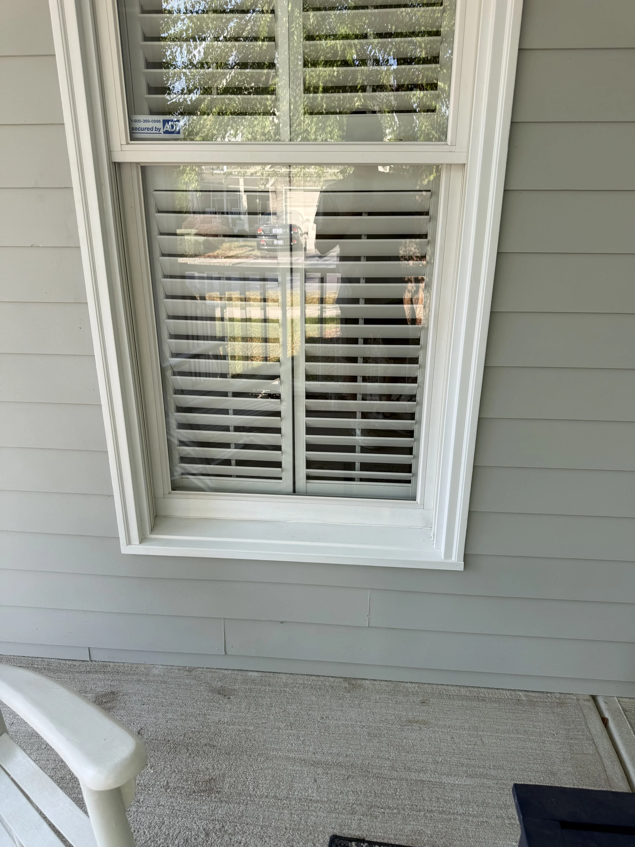 A house window with white shutters on a beige exterior wall, reflecting the outdoors including trees, a car, and a person in the glass. Window frame and Window have been completely cleaned