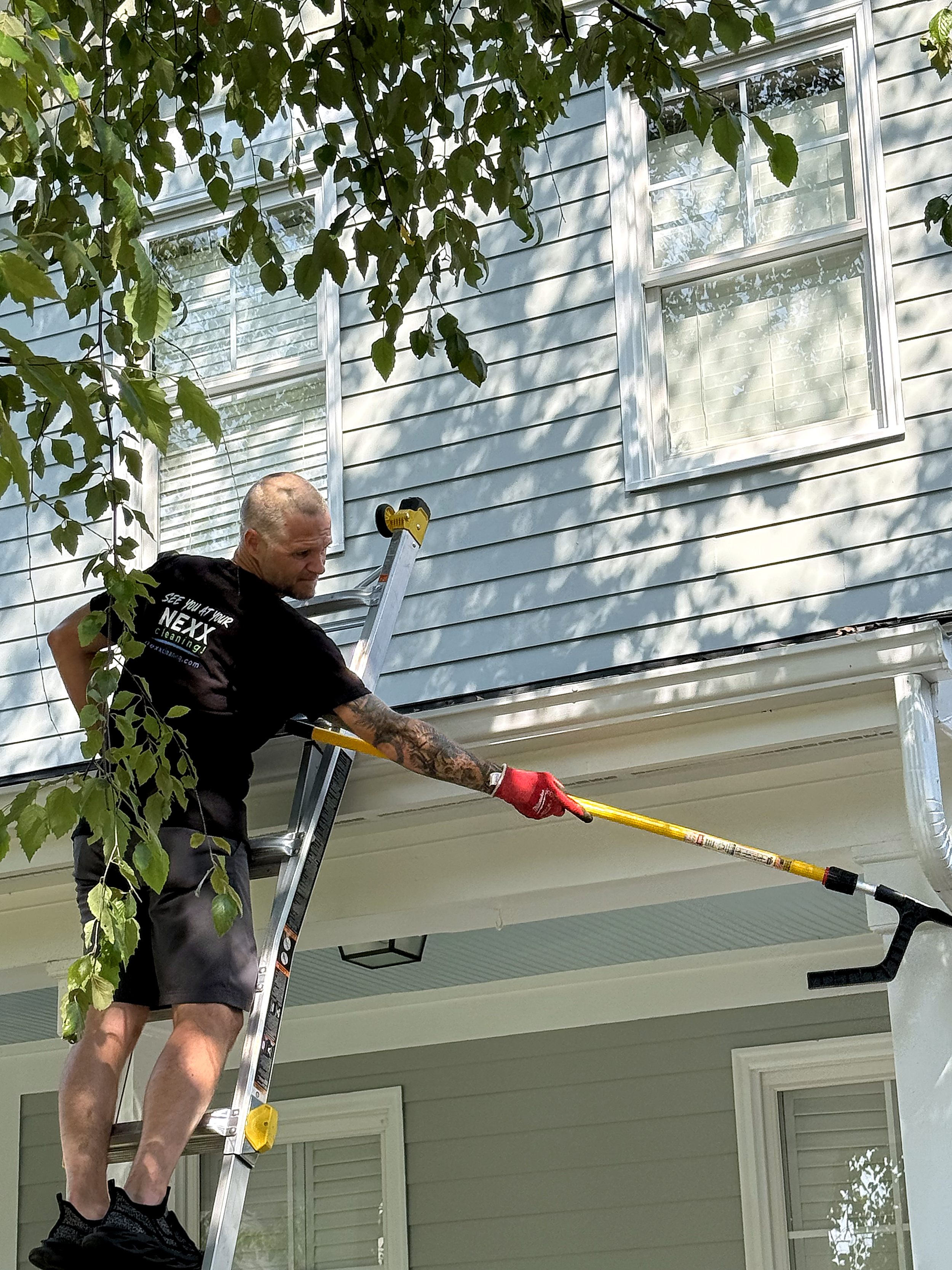 A man standing on a ladder cleaning the exterior of a house with a long-handled paint scraper or cleaning tool. The house has white siding, and there are two windows with white trim. The scene is shaded by tree branches with green leaves.