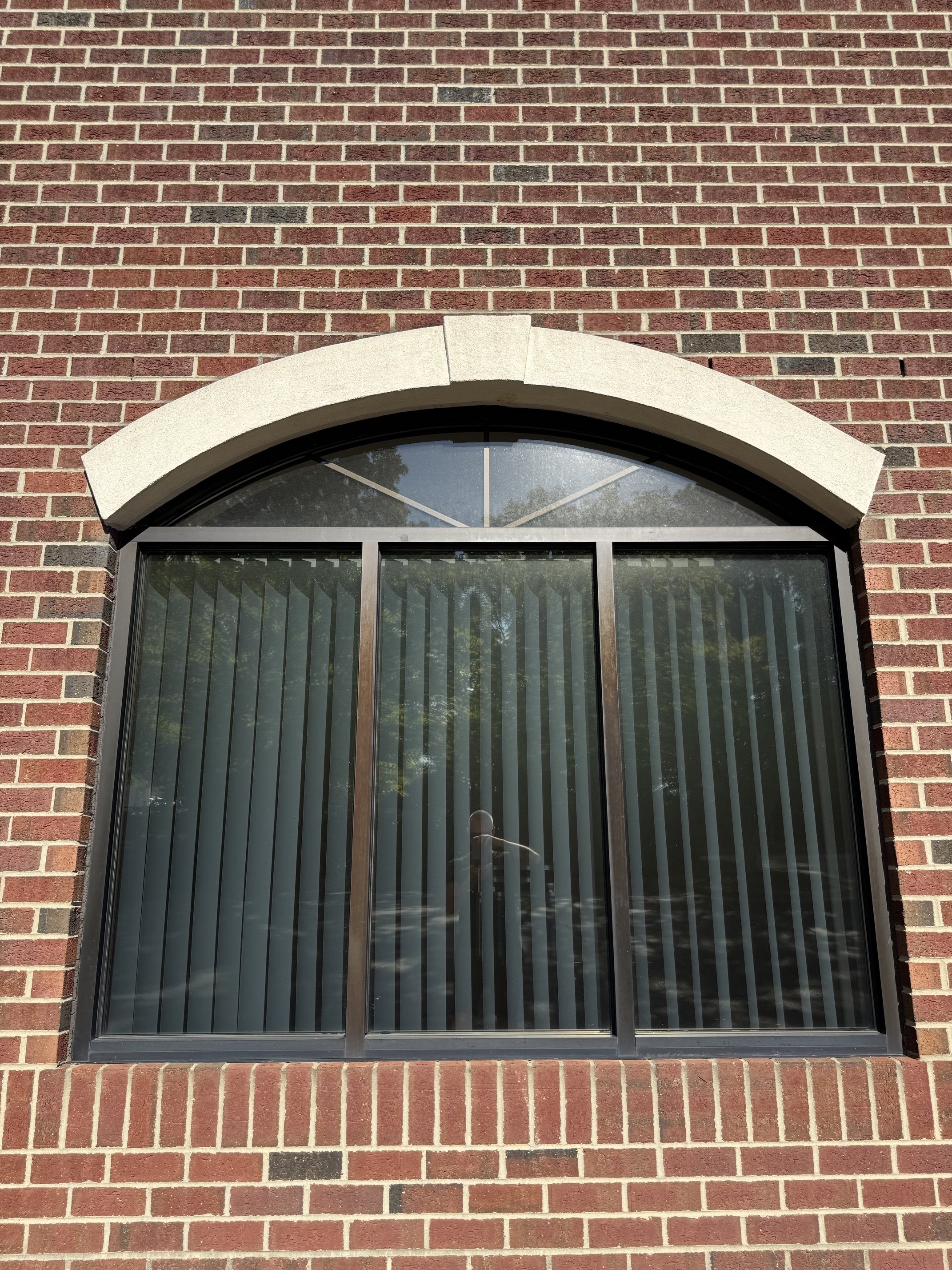 A rectangular window with an arched top set into a brick wall. The window has vertical blinds and a reflection of trees and the sky.