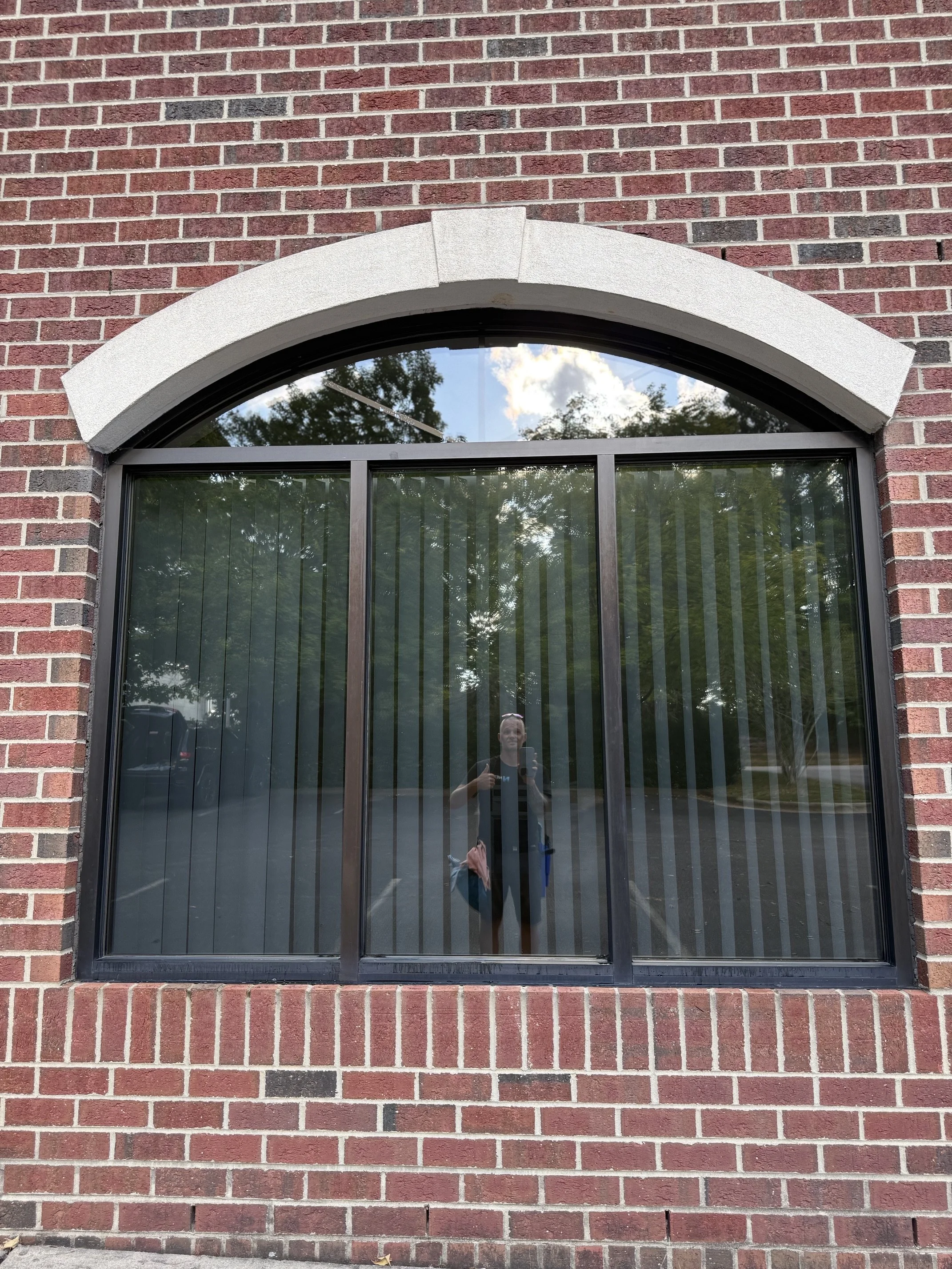 A large reflective window with an arched top, set into a red brick wall, reflecting trees and clouds outside. A person is visible in the reflection taking a photo.