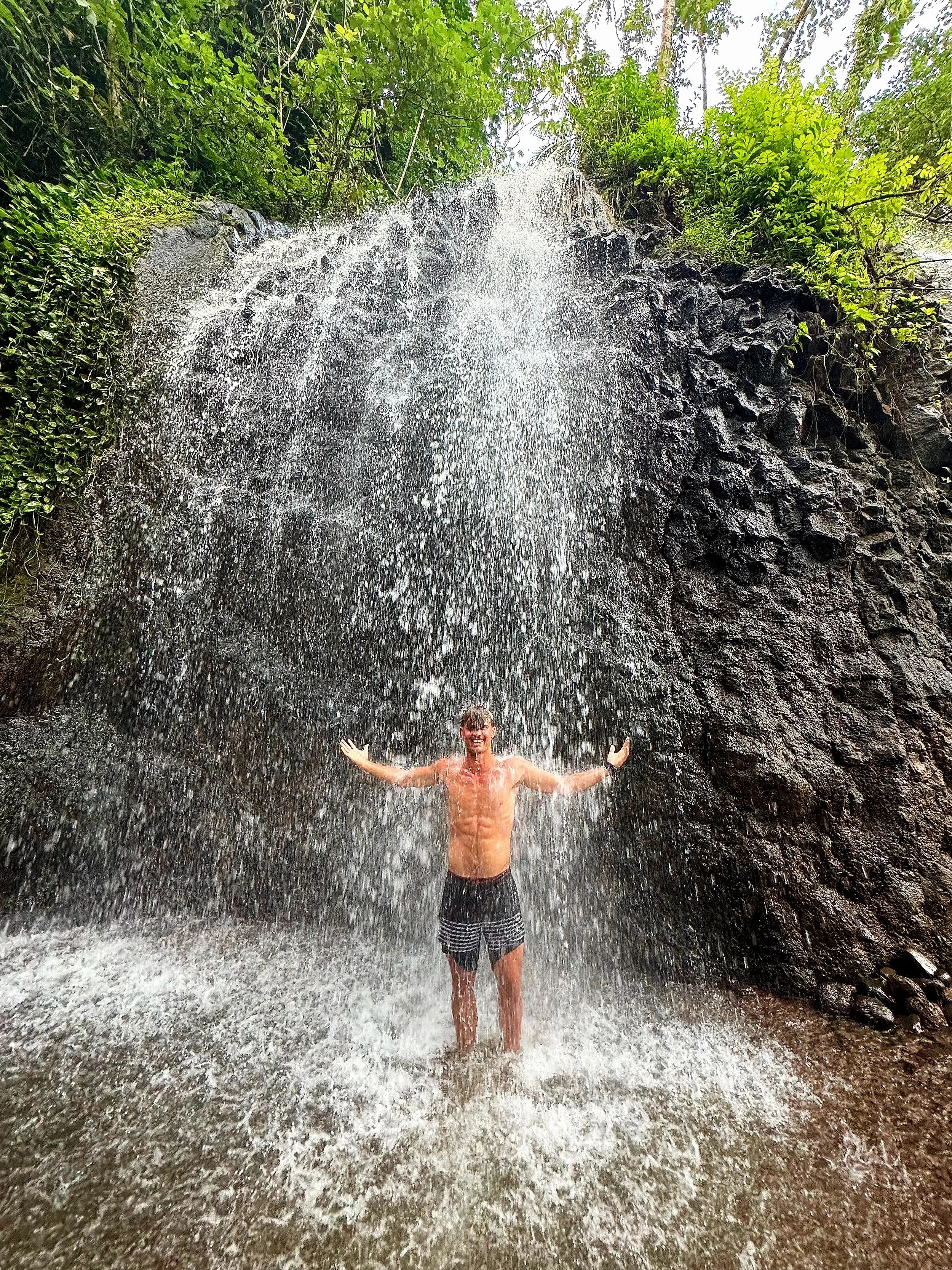 A man standing under a waterfall with water cascading down behind him and lush green foliage surrounding him.