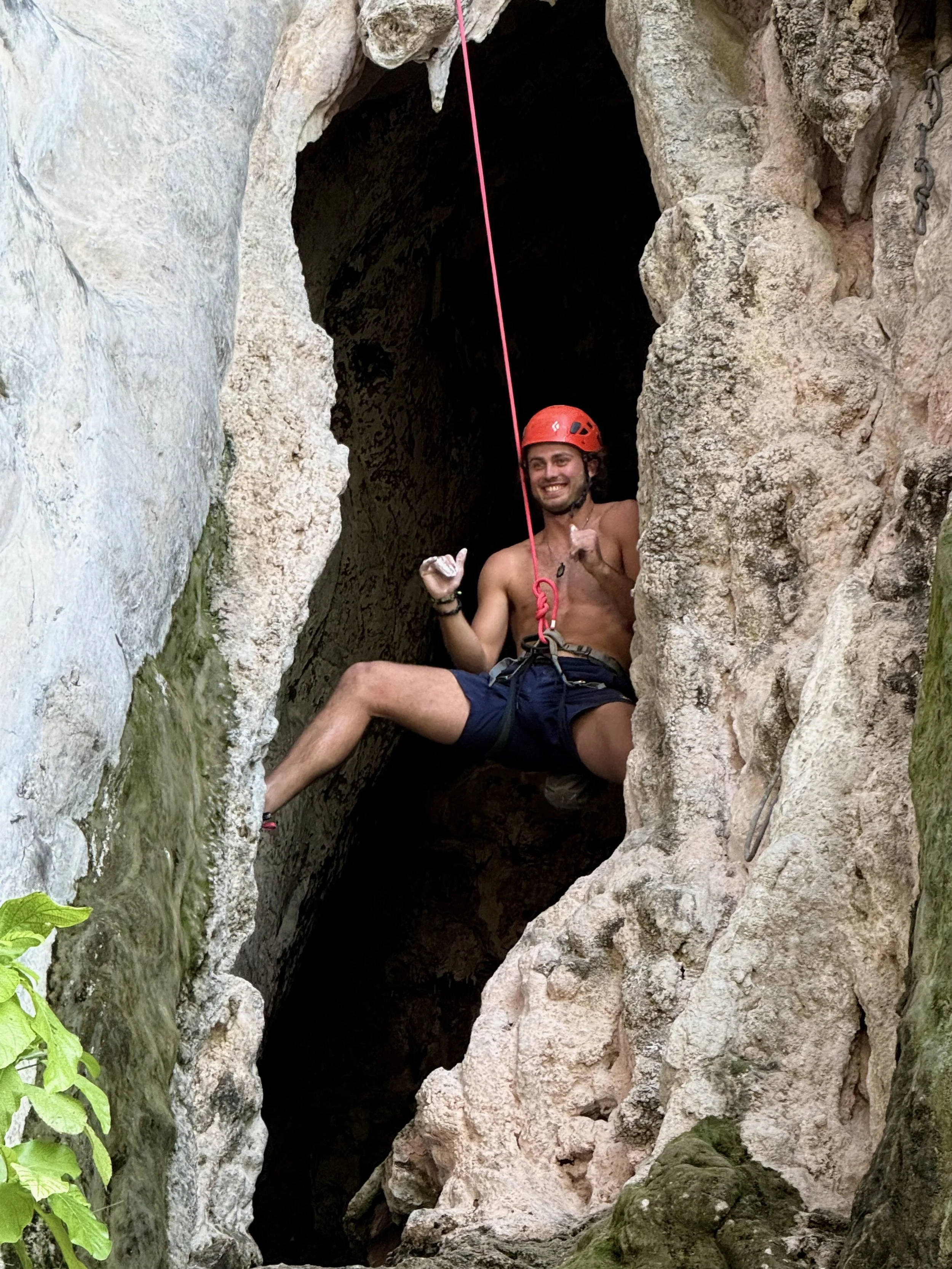 A man with a red helmet, shorts, and a harness sitting in a rocky crevice with a smile, surrounded by natural rock formations.