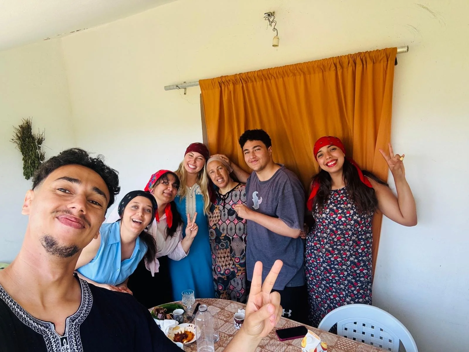 Group of seven people, smiling and posing indoors, some making peace signs. They are gathered around a table with food and drinks, with a yellow curtain and plain wall in the background.
