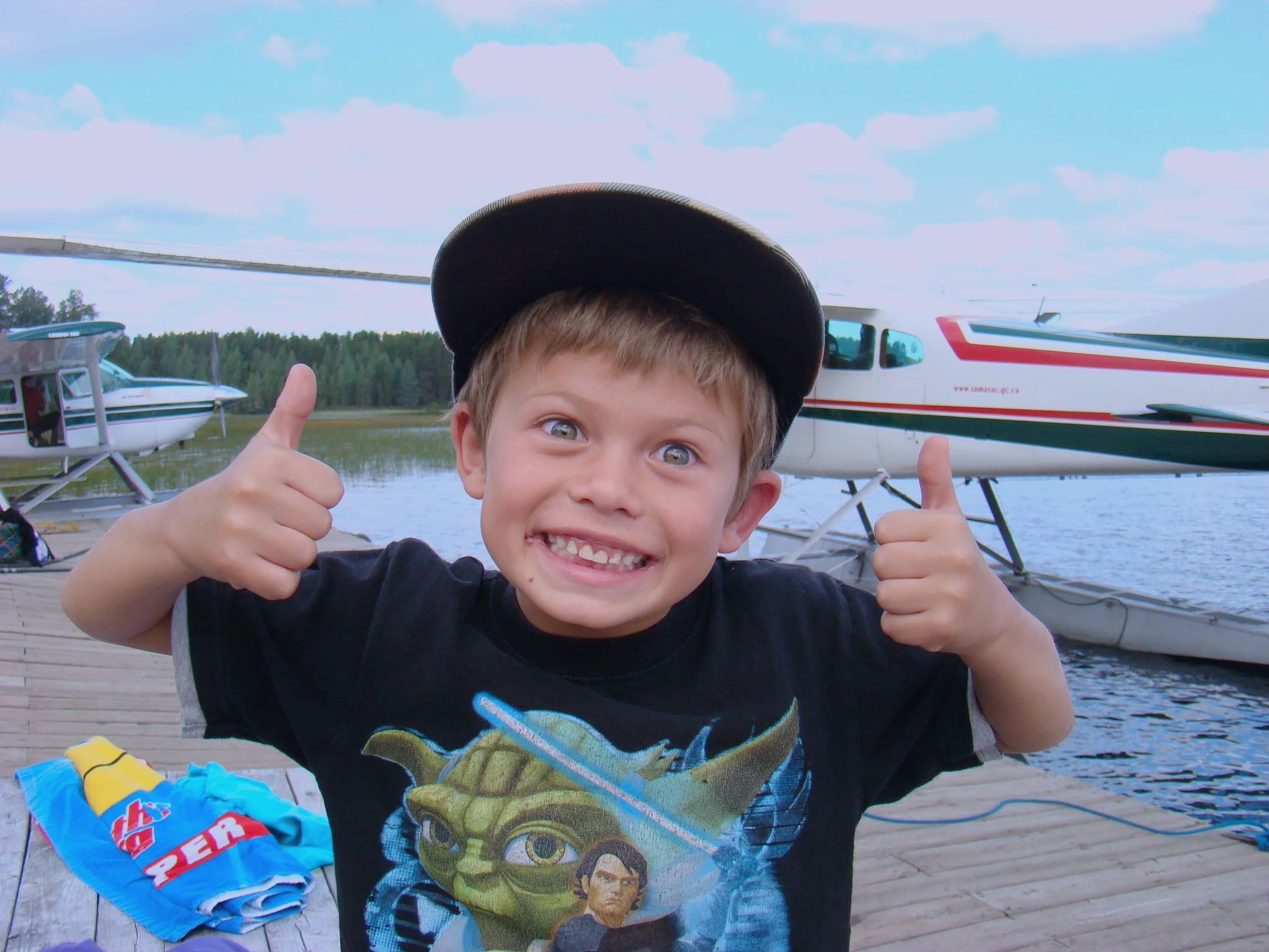 A young boy with short hair and blue eyes wearing a black Batman t-shirt and a baseball cap, smiling and giving a thumbs-up gesture. Behind him, there are small airplanes and boating equipment on a wooden dock near a body of water, with trees and a cloudy sky in the background.