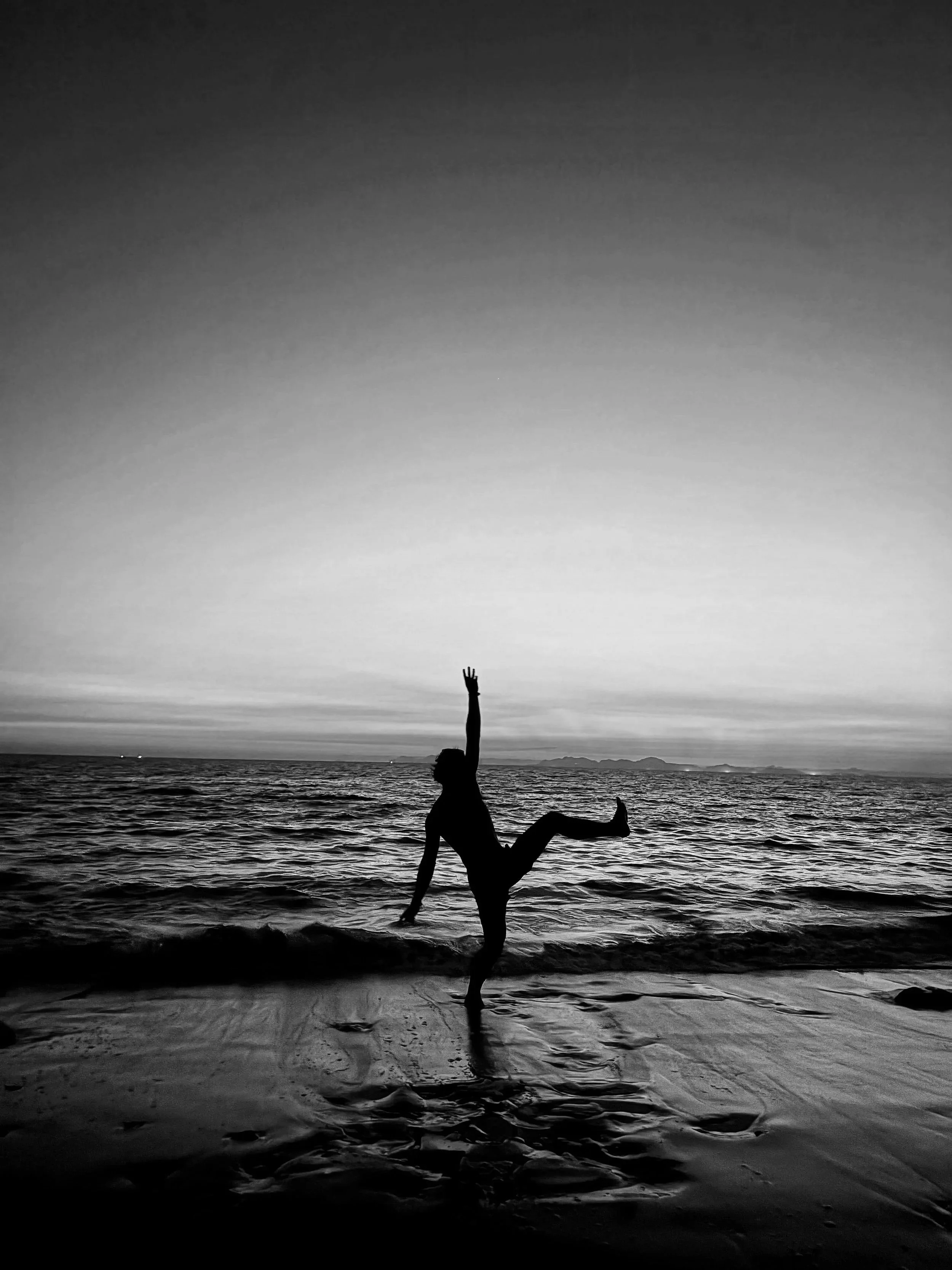 Person doing a handstand on the beach at sunset, with the ocean and distant islands in the background, black and white photo.