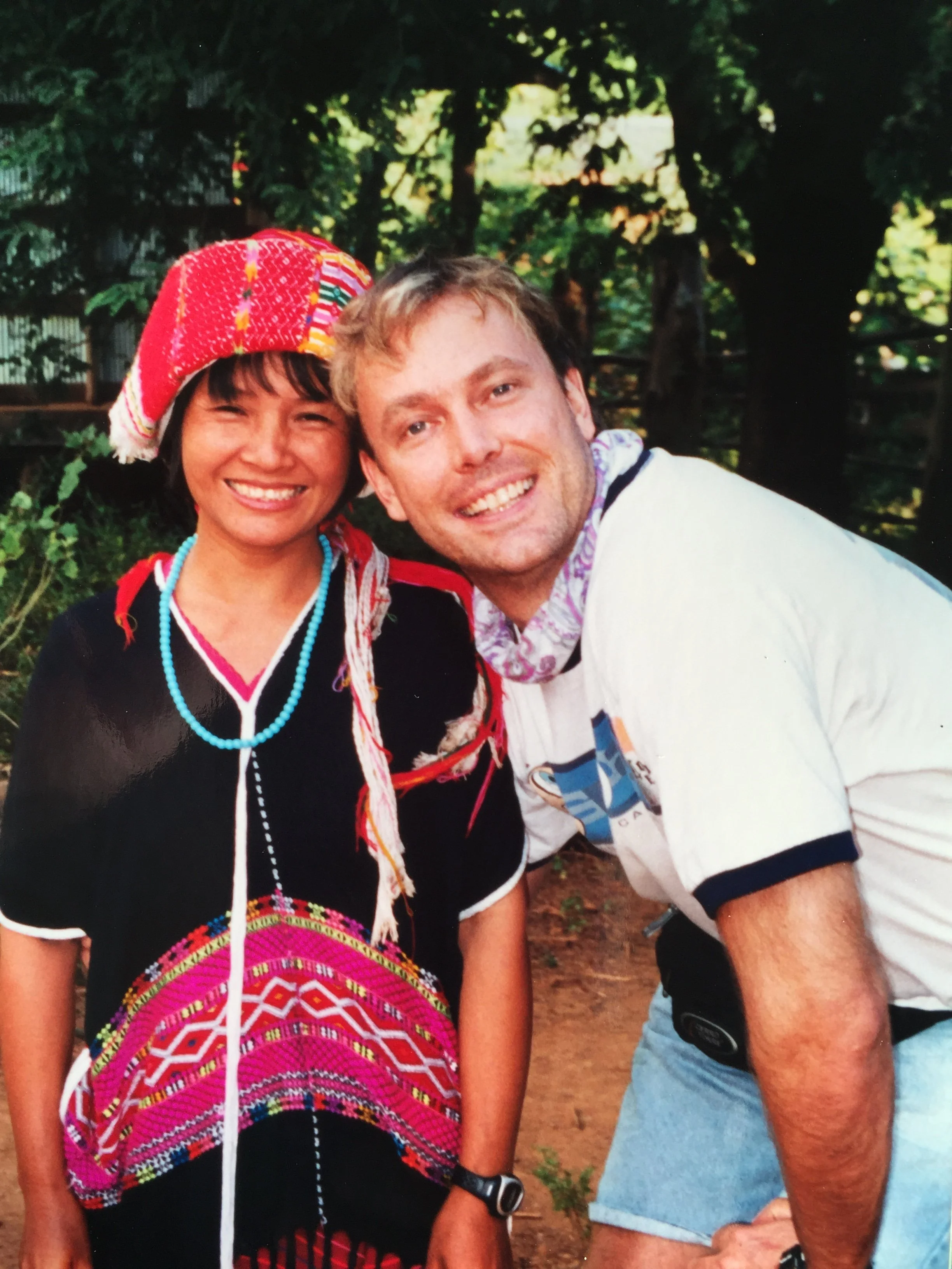 A smiling woman wearing traditional clothing including a colorful hat and necklaces, with a man standing next to her in casual clothing, outdoors in a wooded area.