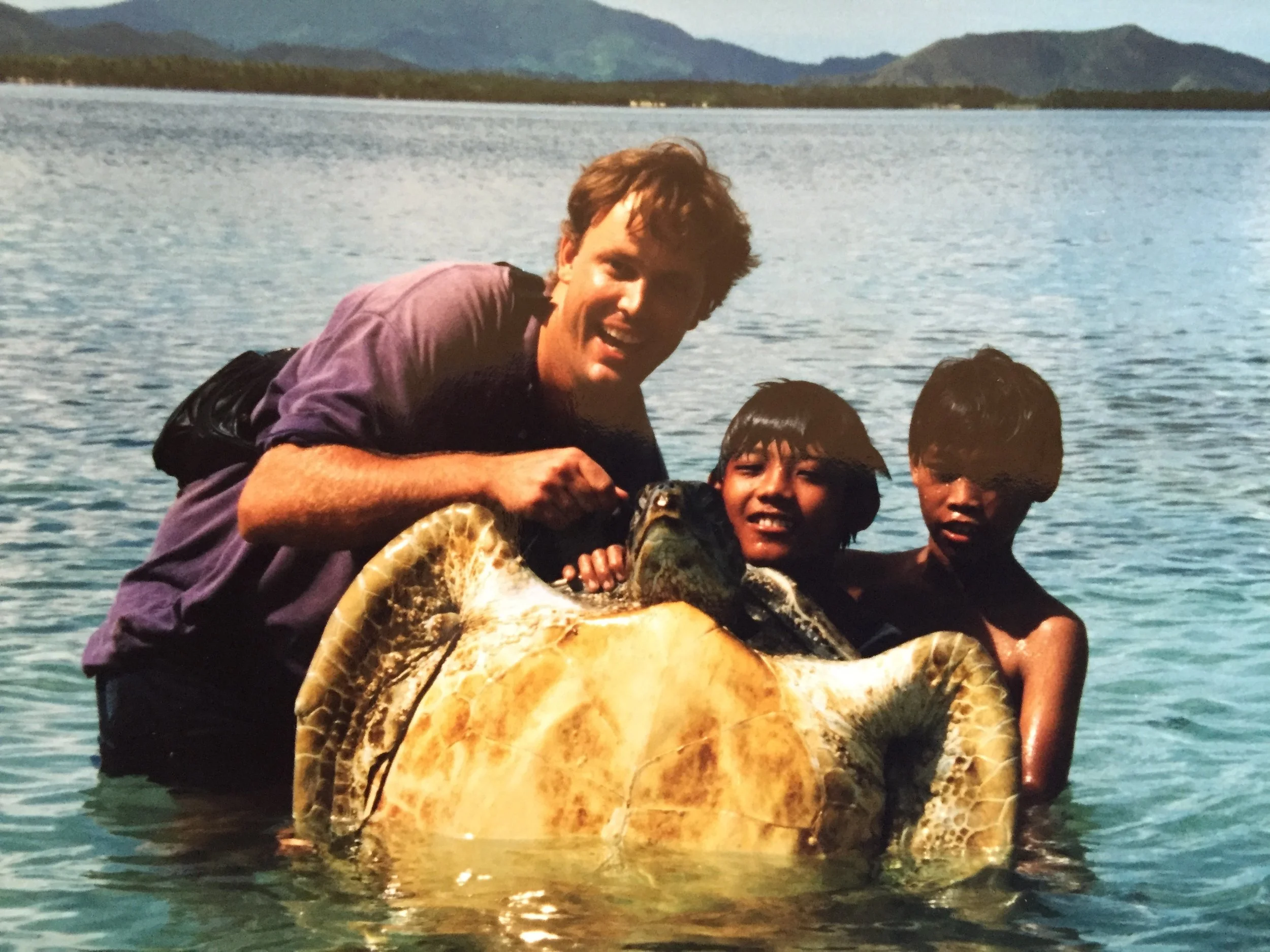 A man and two boys in the water holding a large fish, with mountains and water in the background.