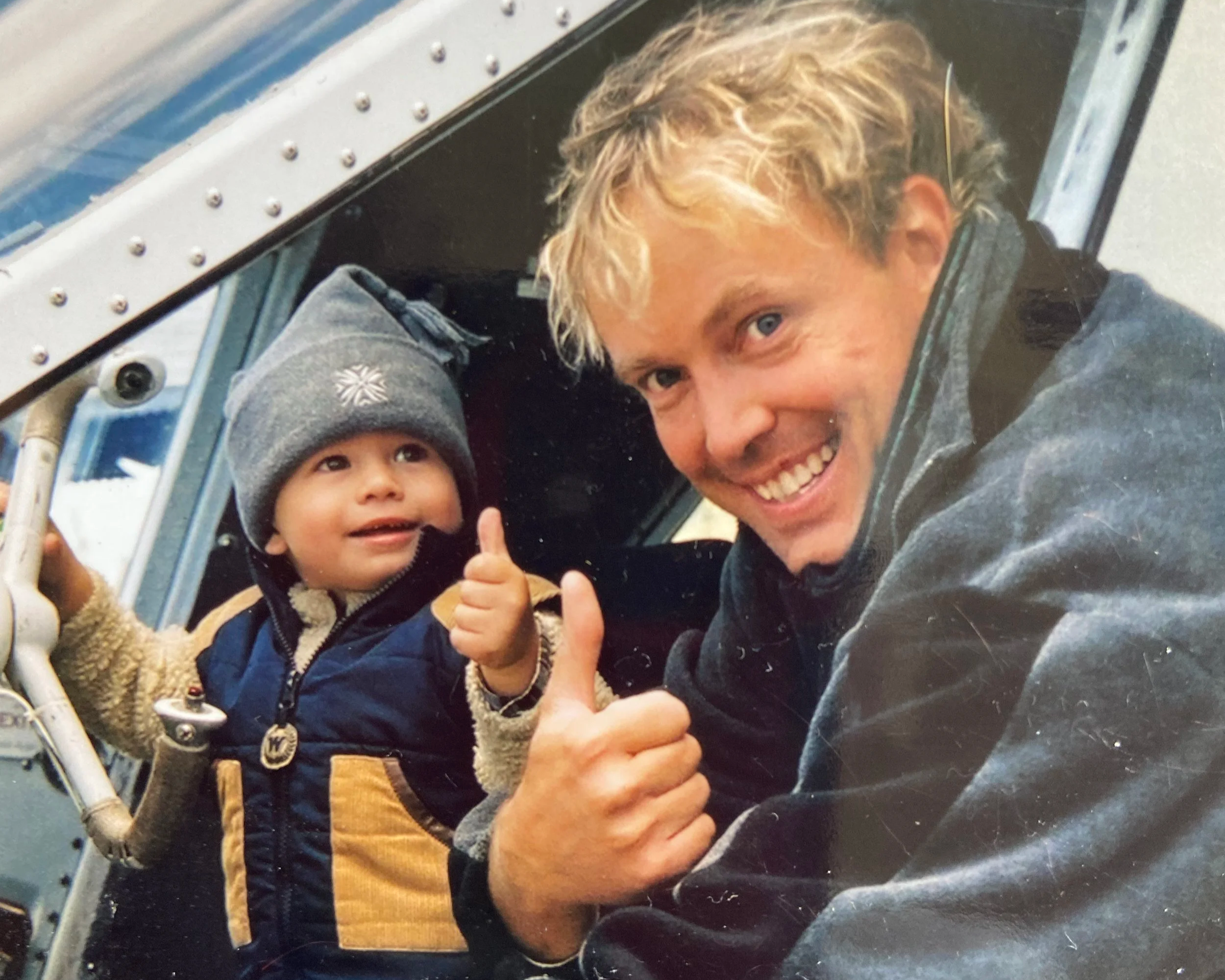 A smiling man and a young child inside an aircraft, both giving a thumbs-up gesture.
