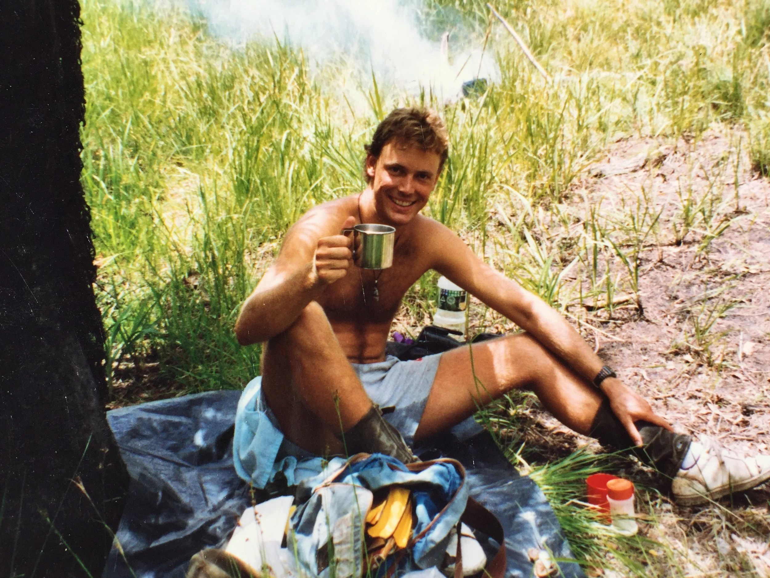 A shirtless man with short brown hair smiling, sitting outdoors in a grassy area next to a small body of water, holding a metal cup.
