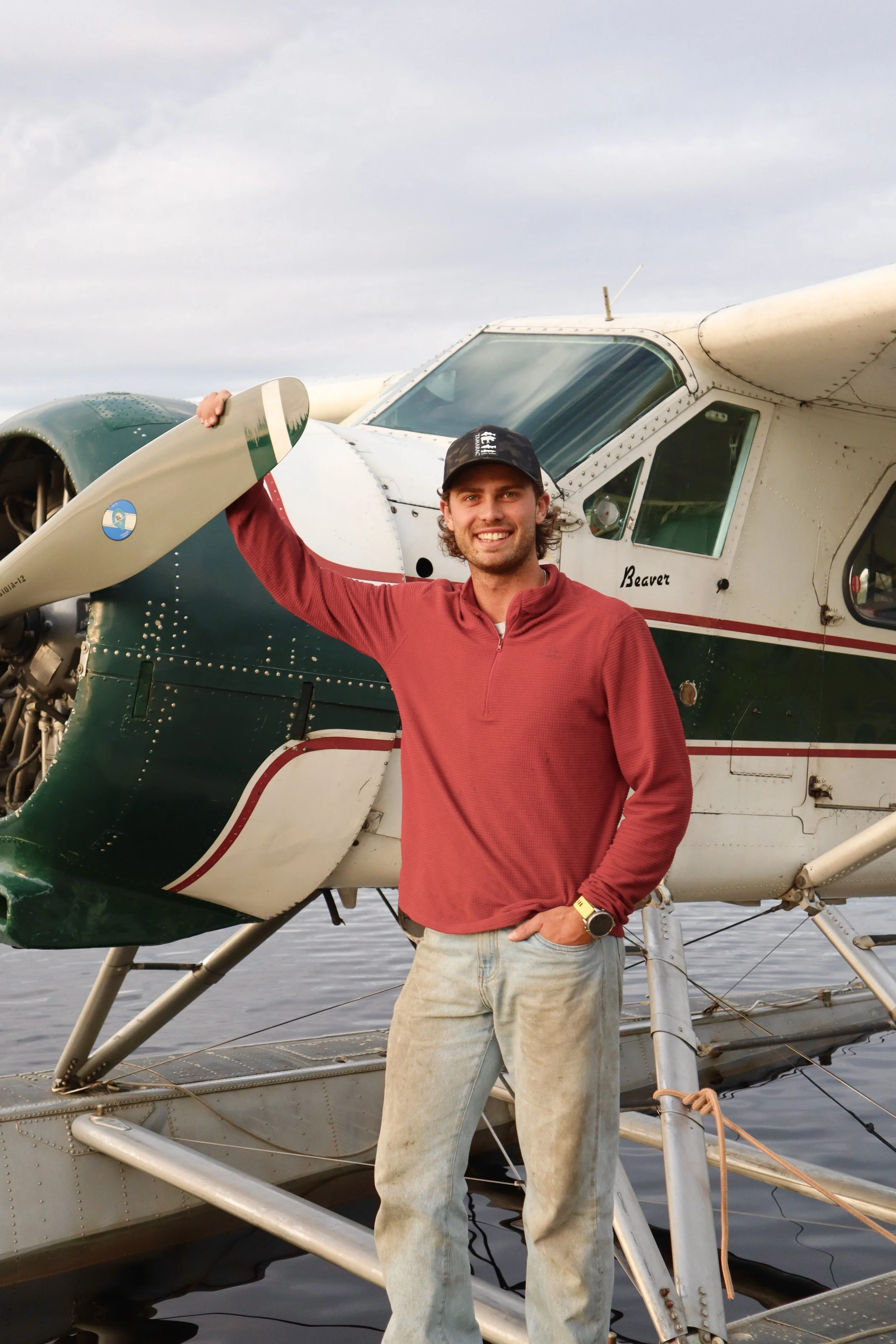 A young man standing in front of a small helicopter, smiling at the camera, with water in the background.