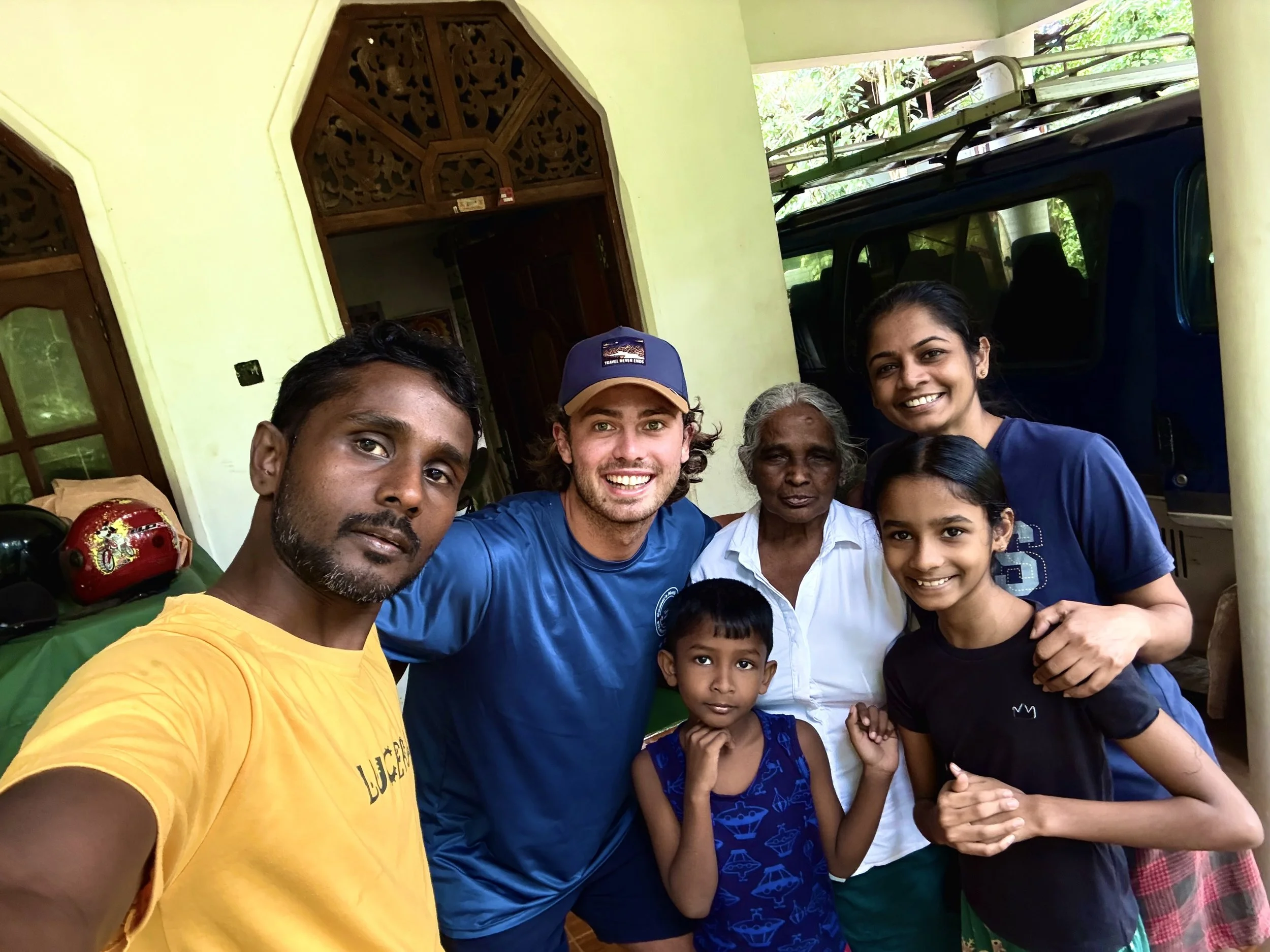 A group of people, including men, women, and children, smiling and posing for a selfie in front of a house with green walls and a large vehicle in the background.