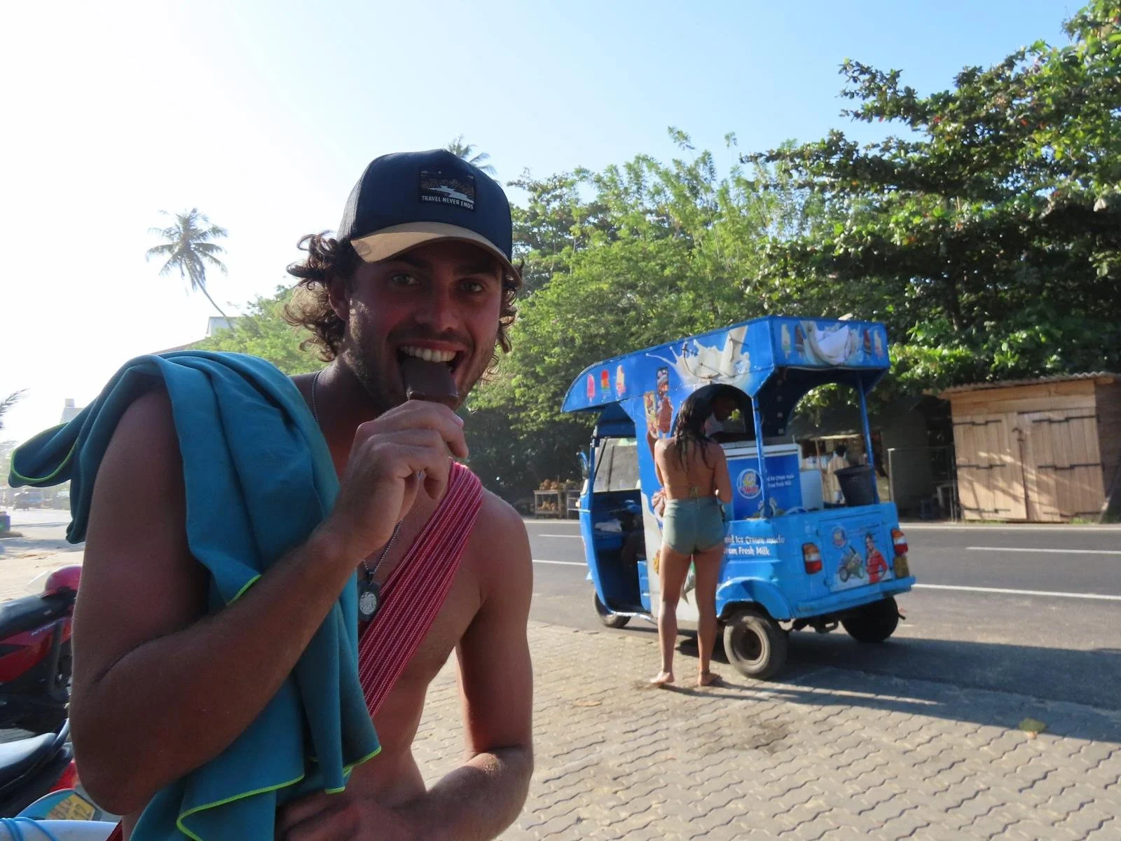 A man with curly hair, wearing a cap and a towel over his shoulder, smiling and licking an ice cream, standing near a blue ice cream truck on a sunny day with greenery in the background.