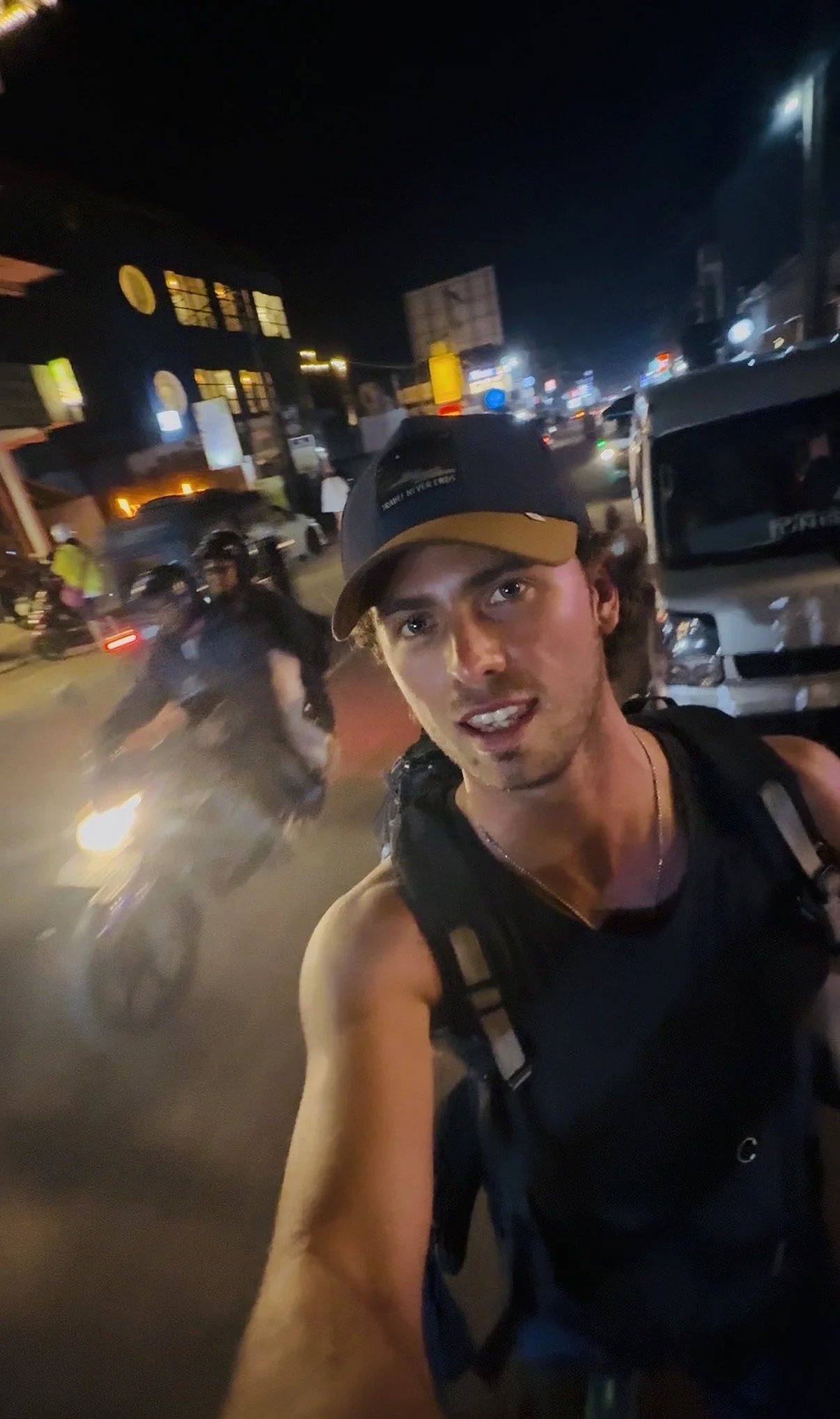 A young man taking a selfie at night on a busy street with cars and motorcycles passing by in the background.