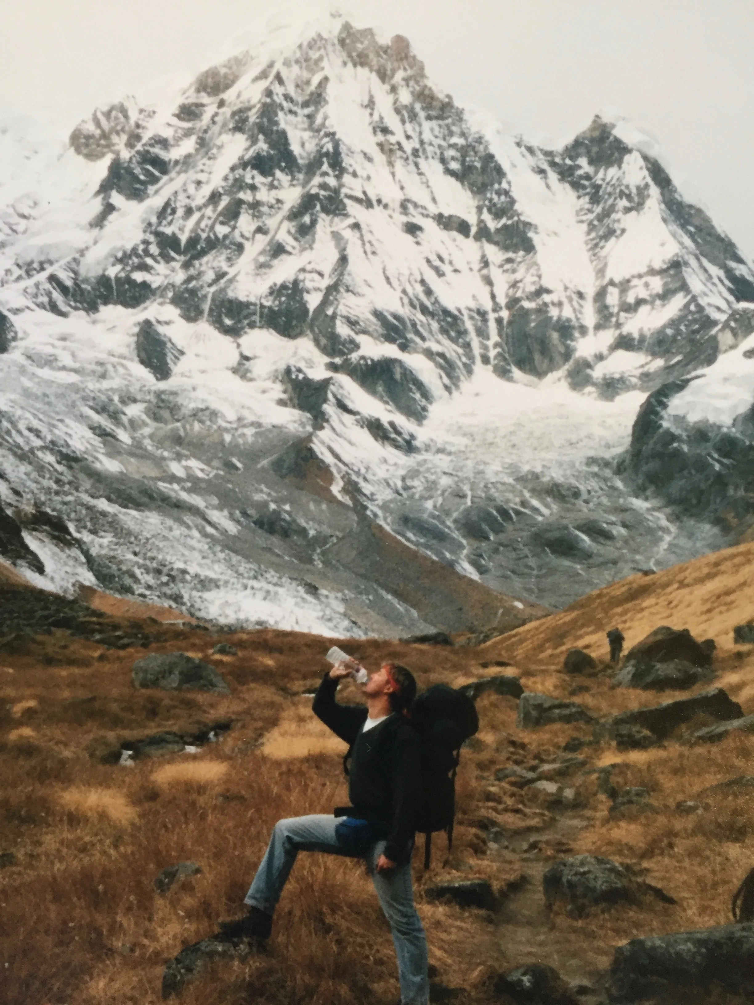 Person sitting on a rock in front of a mountaintop with snow, drinking from a water bottle.