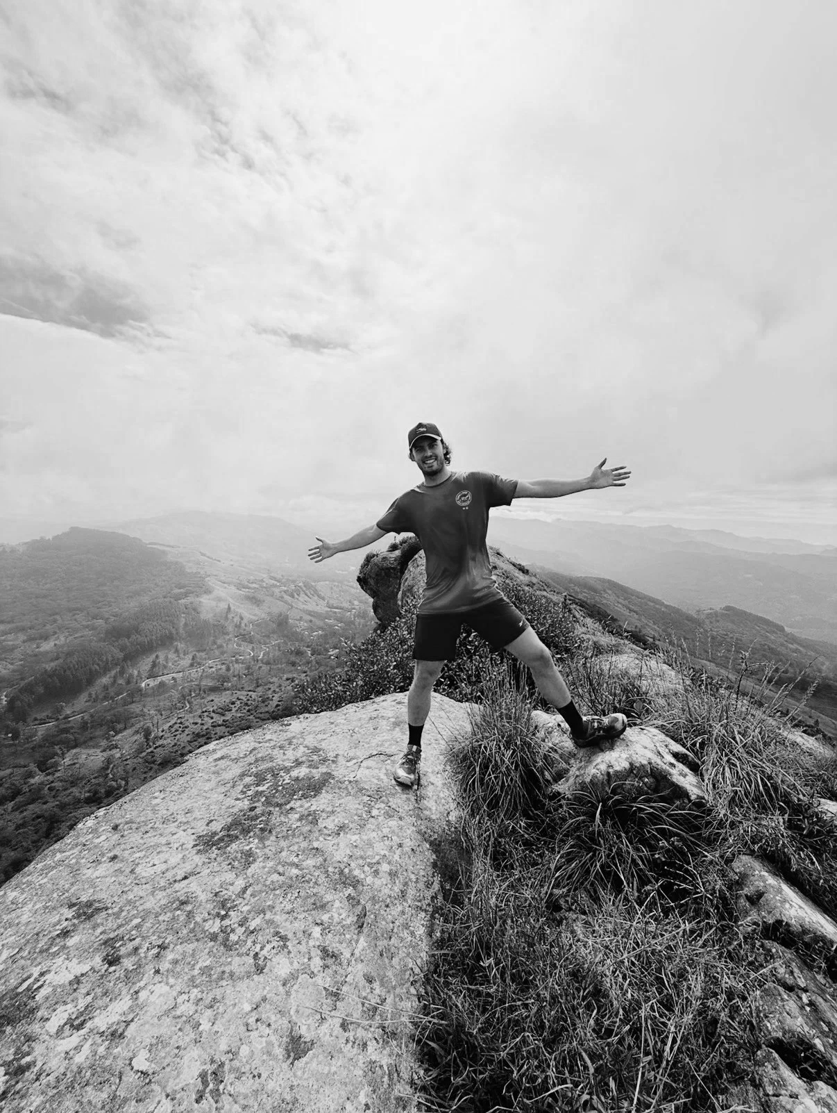 A man standing on a rock on a mountain summit with arms outstretched, overlooking a vast landscape of hills and valleys under a cloudy sky.