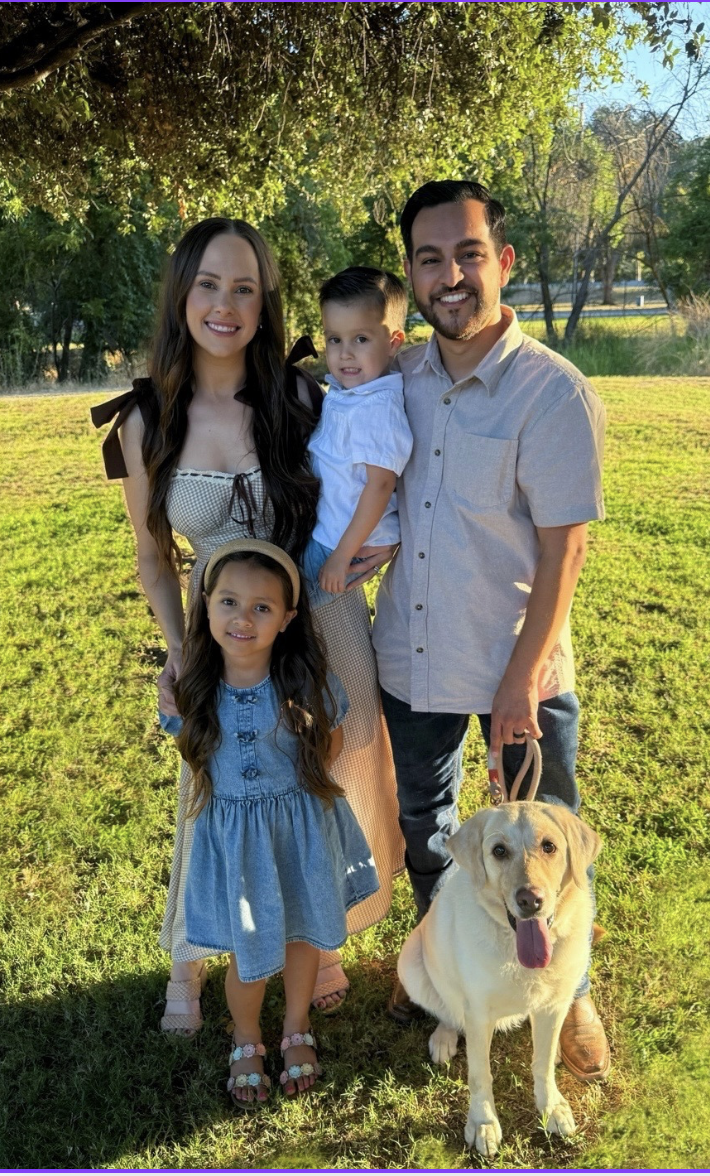 A family of four, including a man, a woman, a young girl, and a young boy, standing outdoors on a sunny day, along with a yellow Labrador Retriever dog. They are smiling and posing under a large tree.