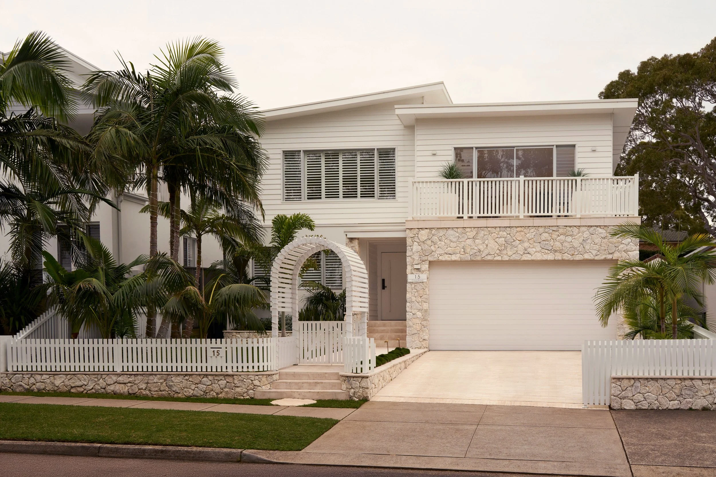 A modern white house with a stone facade on the lower level, surrounded by palm trees and a white picket fence, with an open driveway and a well-maintained lawn.