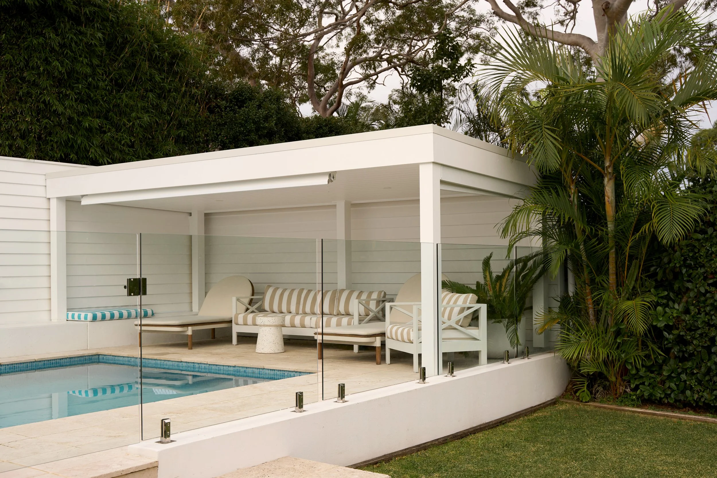 A modern outdoor patio area with a white structure, striped beige and white outdoor furniture, and a glass pool fence surrounding a swimming pool, with lush green trees and plants in the background.