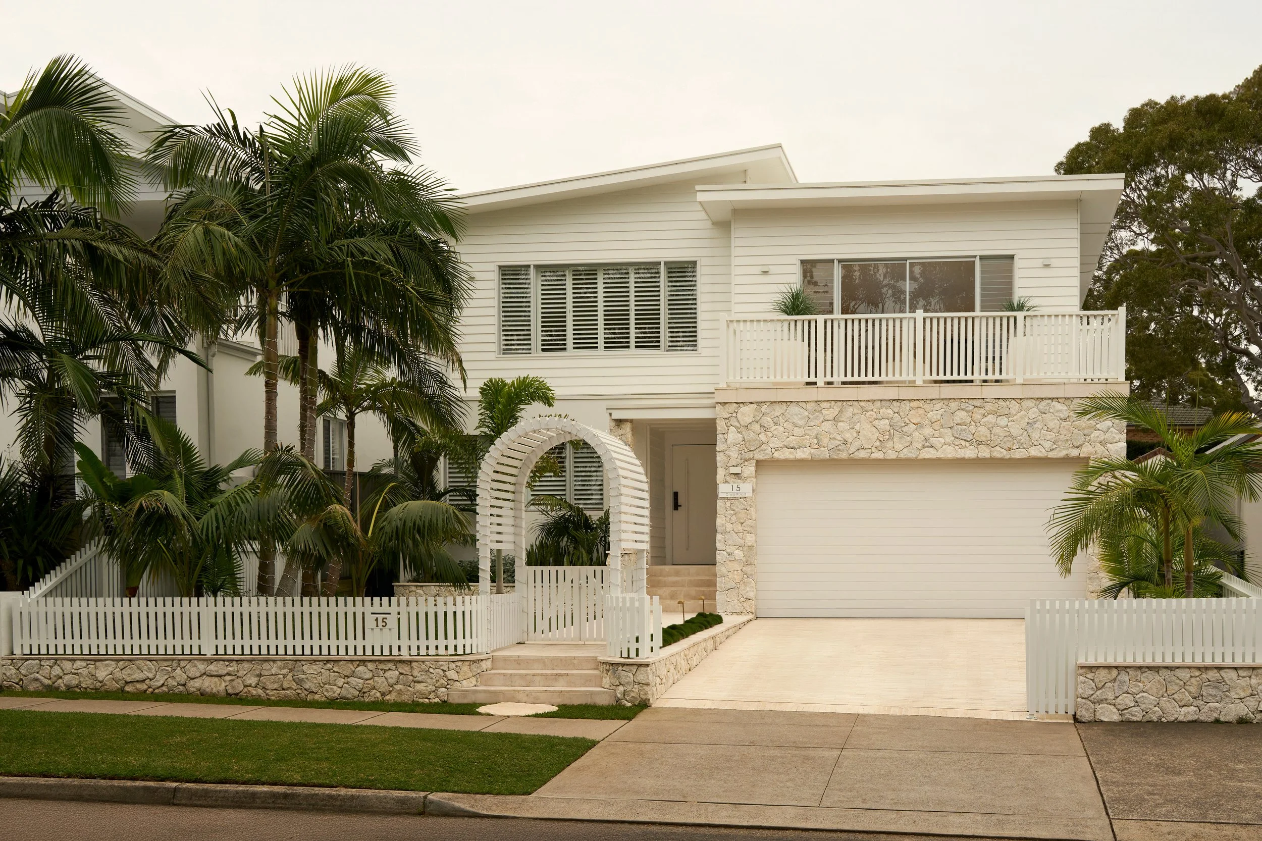 A modern white house with stone accents and a balcony, surrounded by lush palm trees and a white picket fence.
