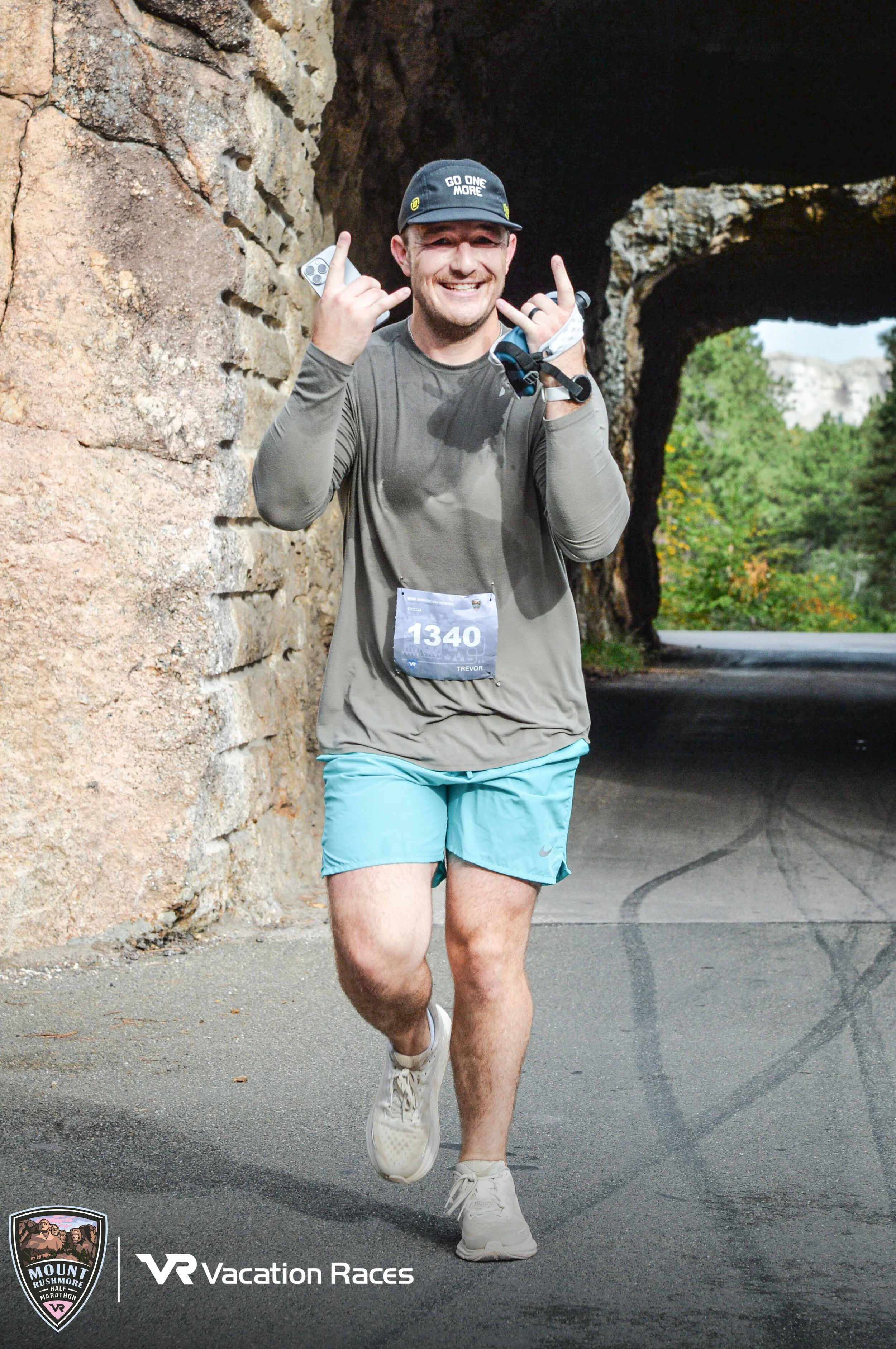 A man smiling and making a rock and roll hand gesture, running outdoors near rock formations and trees. He is wearing a baseball cap with the phrase 'Go One More', a gray long-sleeve shirt, turquoise shorts, and running shoes, with a race bib numbered 1340. The bottom left has a logo and text for Mount Rushmore Half Marathon, and the bottom right says VR Vacation Races.